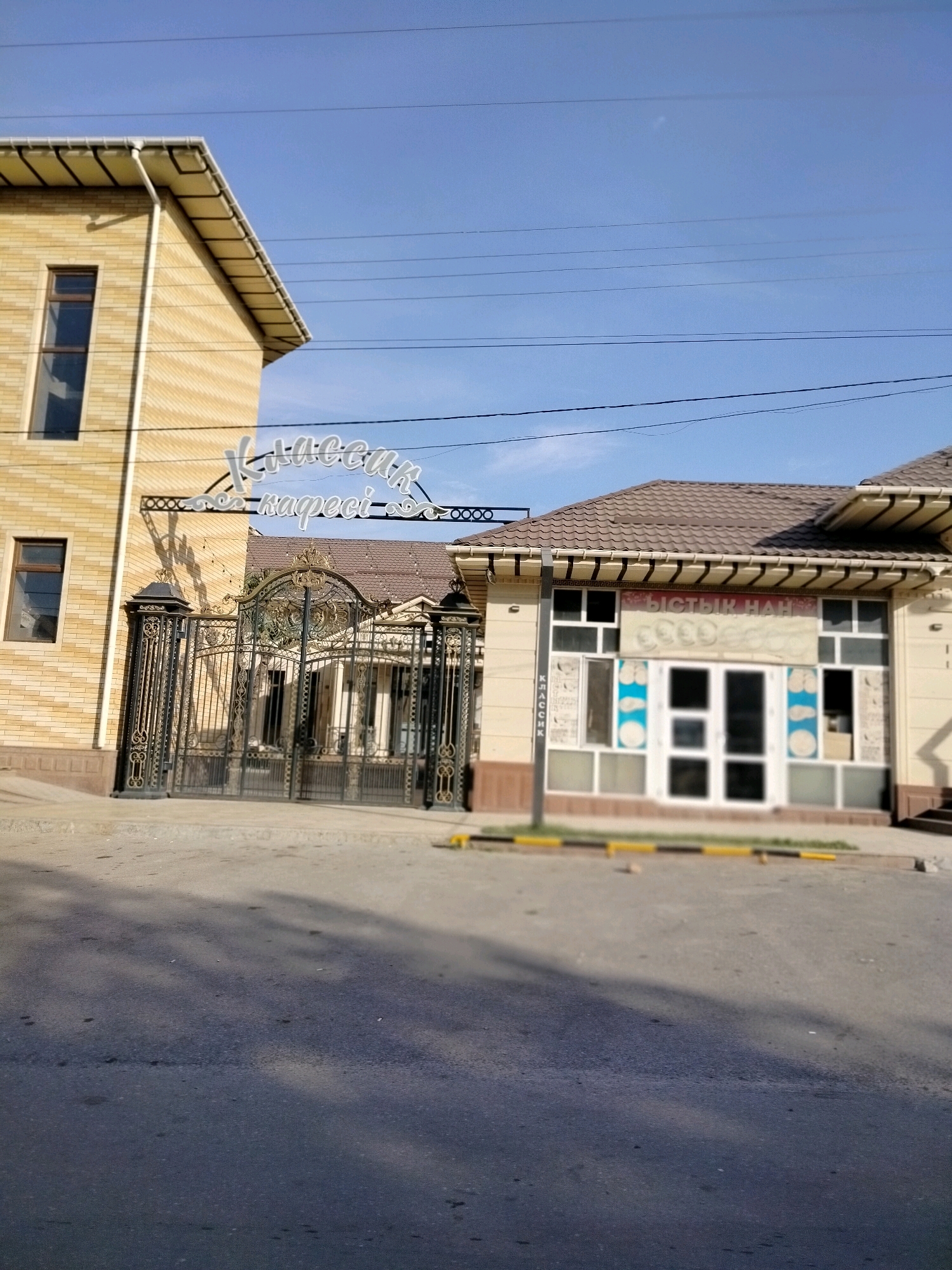 A clear daytime, eye-level outdoor shot shows the entrance to a commercial establishment. The establishment has two buildings, a larger yellow brick one on the left and a smaller beige one on the right with a brown tiled roof. A decorative black wrought-iron gate is set between the two buildings, and above the gate, a white sign with curved, stylized lettering reads "Классик кафесі" (Klassik kafesi). The beige building on the right has a sign above its windows that reads "Ыстык Нан" (Ystyq Nan), which translates to "hot bread." The windows of the beige building are boarded up with white material and feature decorative elements. The ground in front of the establishment is paved with asphalt and a sidewalk separates the street from the building. The sky is clear and blue. No people are visible in the image. The location is identified as Көлкент, Kazakhstan.