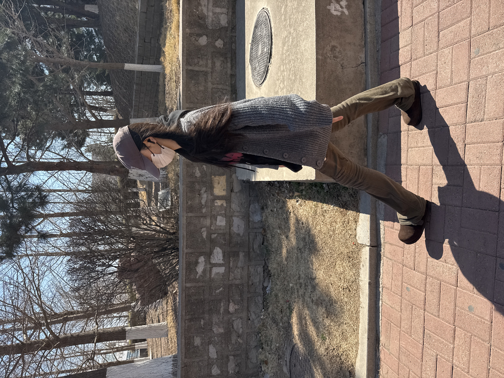 A person stands on a brick pathway in Zhongtun, China. The person wears a gray cap and a white face mask, a gray cardigan, and brown pants with brown shoes. To the left of the pathway is a strip of unkempt lawn bordering a stone wall. A bare tree stands behind the wall, with a glimpse of blue sky in the background. To the right of the pathway, a raised concrete platform contains a circular manhole cover. The time of day is daytime, and the scene appears to be outdoors.