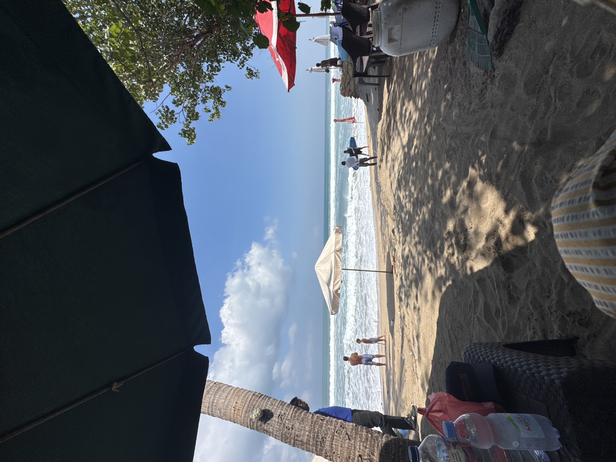 The image depicts a sunny day at Kuta Beach, Indonesia. In the foreground, a black umbrella casts a shadow over the sandy beach. To the left, a person stands near a palm tree, partially obscured. Further inland, a beige umbrella shades a patch of sand. Two people, an adult and a child, are visible near the water's edge. The ocean displays moderate waves under a clear blue sky with scattered clouds. To the right, red and white umbrellas are set up near some chairs and tables. Several people are seen near the water, with some appearing to carry surfboards. The scene is a typical beach setting with elements of leisure and activity.