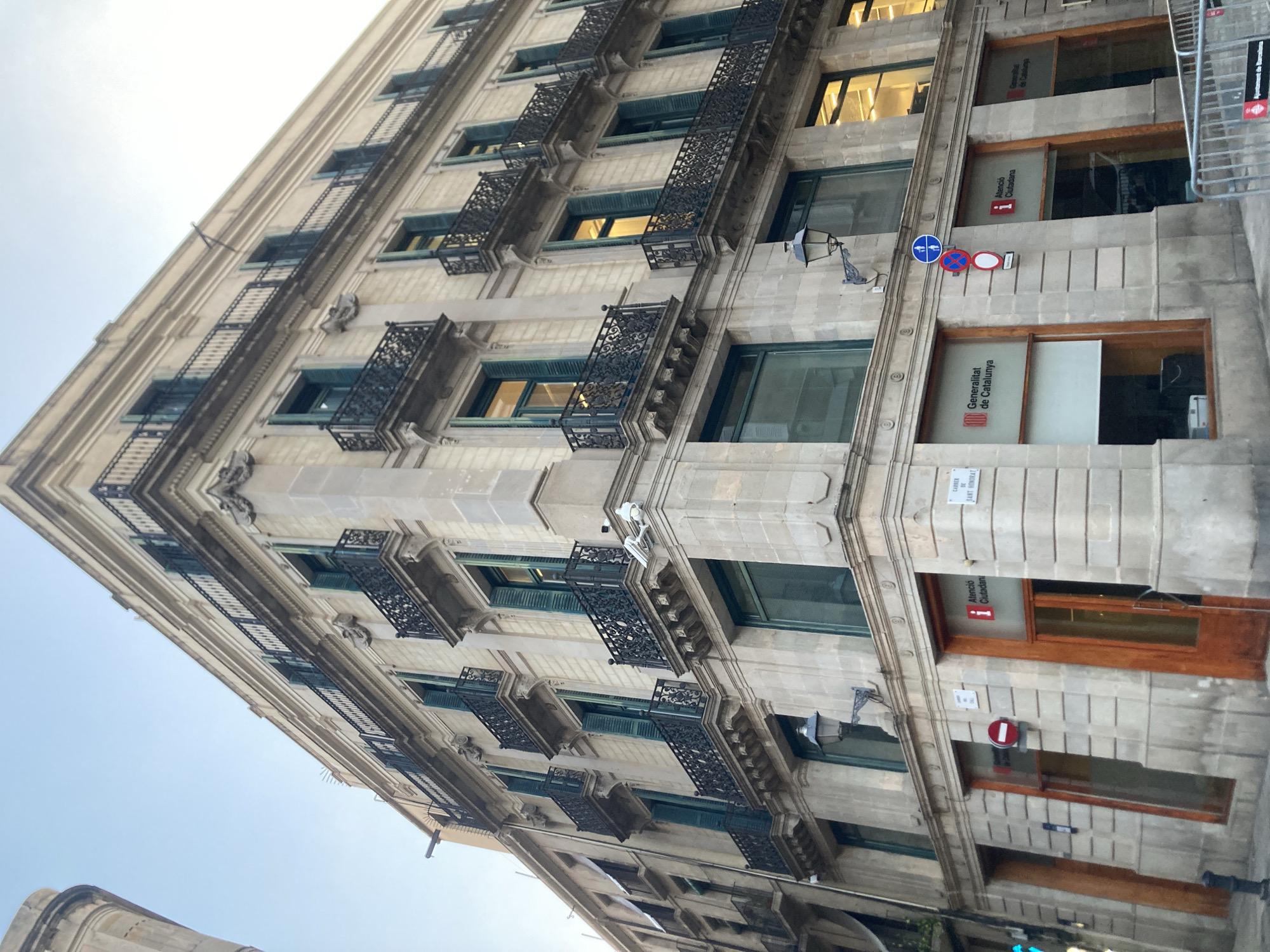 The image depicts a corner building in Barcelona, Spain, featuring traditional architecture with stone facade, balconies with ornate iron railings, and green shutters. The building has multiple stories, with shops or offices on the ground floor. One of the shops has signage that reads "Generalitat de Catalunya" and "Atenció Ciutadana," indicating it is a citizen services office of the Catalan government. There are traffic signs visible, including a pedestrian crossing sign and a circular red and white sign. A security camera is mounted on the corner of the building. The sky is clear and blue, suggesting daytime.