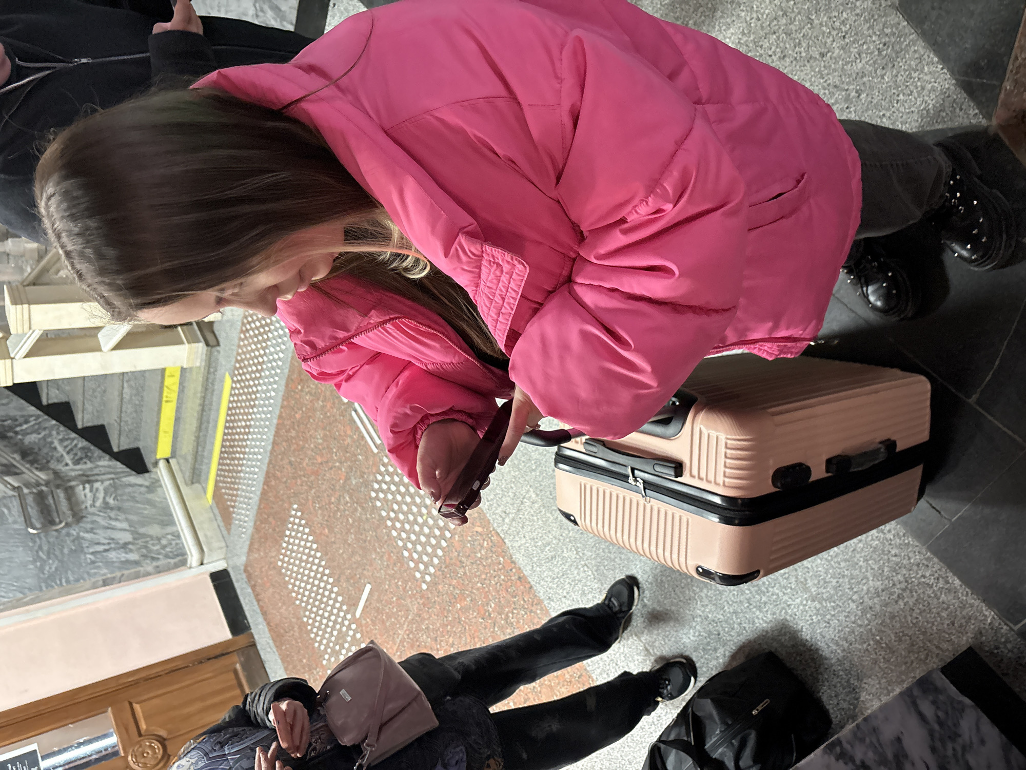 A medium, eye-level shot depicts a young woman in a pink puffy jacket standing on a patterned floor, pulling a light-colored suitcase. The woman has long brown hair and is looking down at the suitcase handle in her right hand. She is wearing dark-colored pants and black shoes. To her left, another individual is wearing a dark jacket and pants with black shoes, standing near a pink purse and a black bag. In the background, there's a glimpse of stairs, a wooden door, and patterned flooring. The environment appears to be indoors, likely a building lobby or station. The location is Izhevsk, Russia.