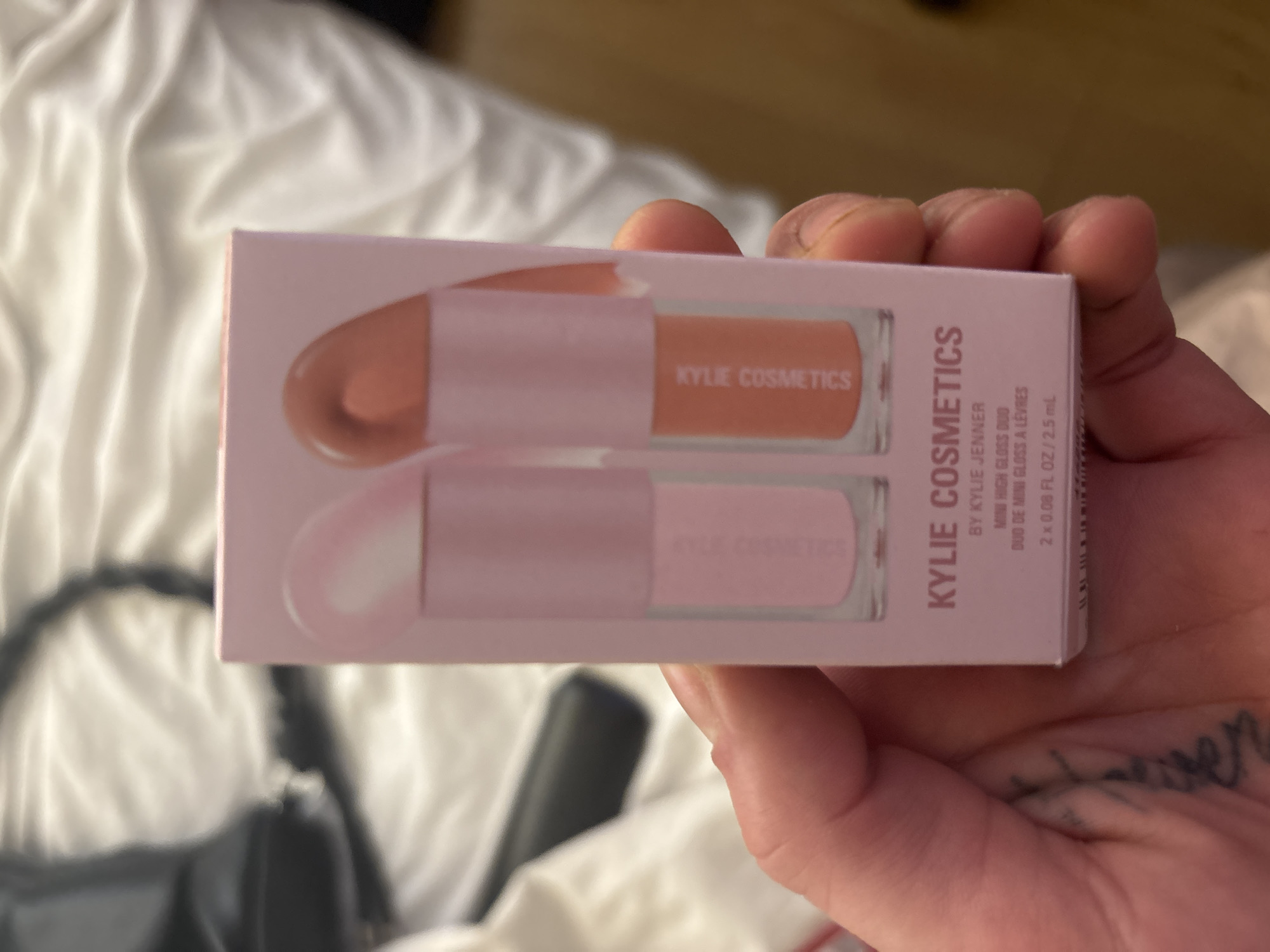 A close-up, eye-level shot features a person's hand holding a small, rectangular light pink box containing two mini lip glosses. The box, branded "KYLIE COSMETICS," has a clear window showcasing the two gloss tubes inside. One tube appears to hold a peachy-nude shade, while the other contains a light pink or clear gloss, both also labeled "KYLIE COSMETICS." The text "BY KYLIE JENNER," "MINI HIGH GLOSS DUO," "DUO DE MINI GLOSS À LÈVRES," and "2 x 0.08 FL OZ / 2.5 mL" is clearly visible on the packaging.
The hand, with visible knuckles and a partial view of a tattoo on the thumb area, holds the product centered in the frame. The background is softly blurred, showing rumpled white or light-colored fabric, likely bedding, suggesting an indoor setting. Some out-of-focus dark objects are vaguely visible in the lower background. The lighting is artificial, making the time of day indeterminate. The image does not contain any visual cues to confirm the specified location of Buenos Aires, Argentina. The activity is the simple presentation or holding of the cosmetic product.