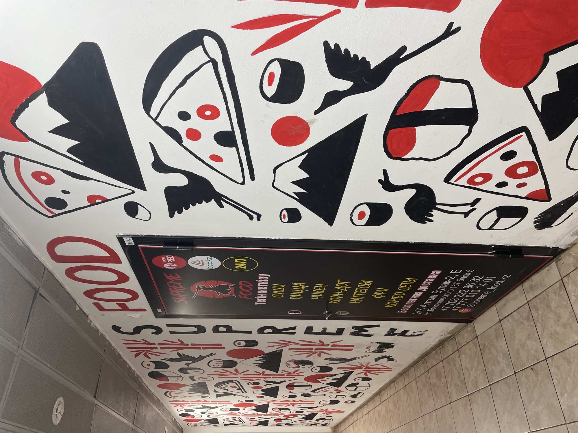 The image depicts a mural on the wall of a building, likely in Almaty, Kazakhstan. The mural features a stylized design with elements of Japanese culture and food, including pizzas, sushi, cranes, and Mount Fuji, rendered in black and red on a white background. The mural is located in a hallway or corridor, with a tiled floor visible in the foreground. To the right of the mural is a black door with a sign that reads "SUPREME FOOD" in red lettering and a logo that appears to be a stylized crane. The door also displays text in Cyrillic, indicating it is likely a business. The presence of Cyrillic text and the mention of Almaty in the prompt suggest the location is within the city. The overall scene is static and appears to be an outdoor or semi-outdoor public space, possibly a walkway or entrance to a commercial establishment.