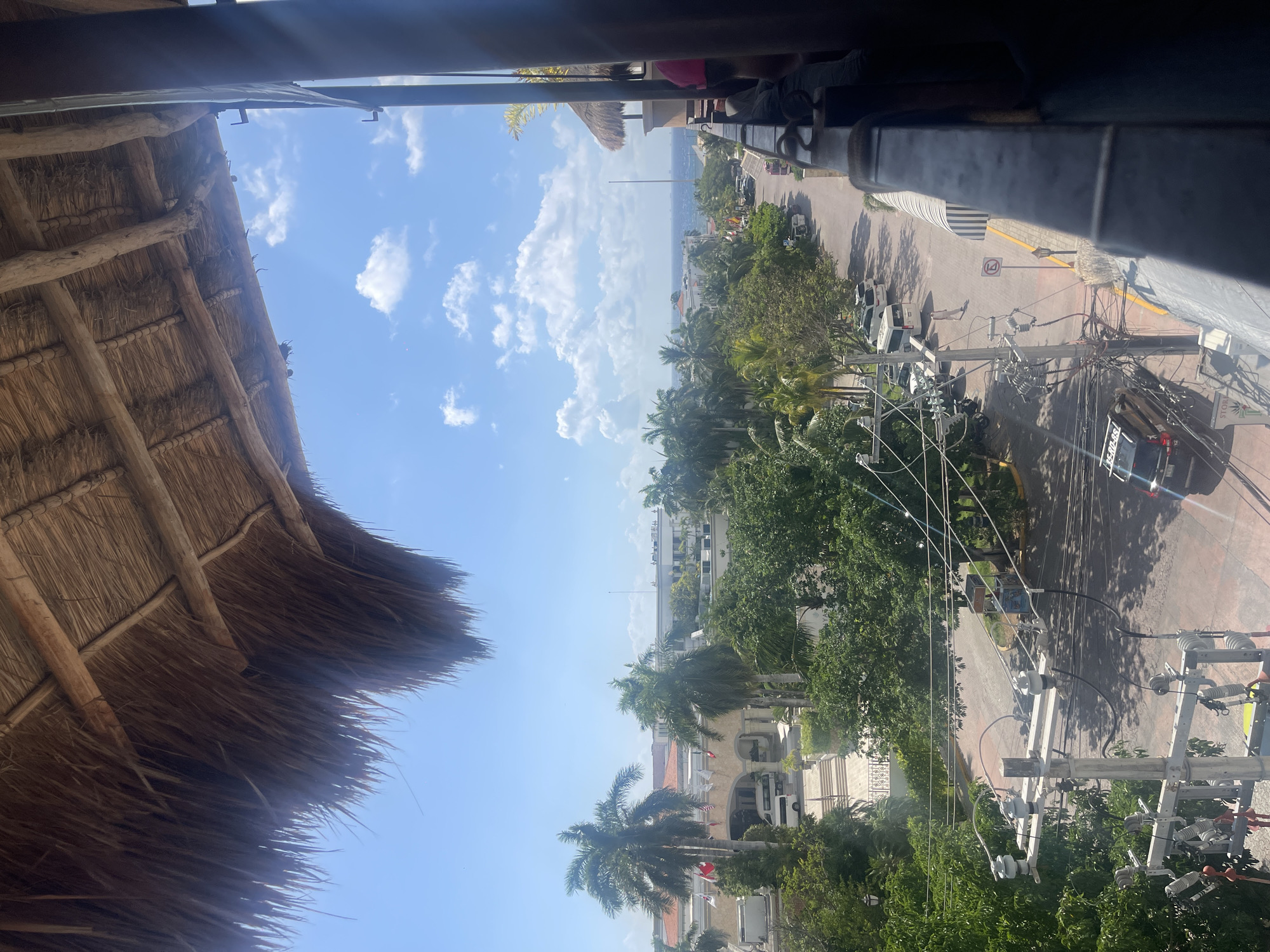 The image captures a view of Playa del Carmen, Mexico, from an elevated position, possibly a balcony or rooftop, revealing a mix of urban infrastructure and natural landscape. A thatched roof structure occupies the left foreground, contrasting with the built environment beyond. A street stretches out, lined with trees and buildings, and dotted with vehicles. Power lines are visible, crisscrossing in the middle ground. The sky is partially visible, displaying blue with scattered white clouds. The sea is glimpsed in the distance.