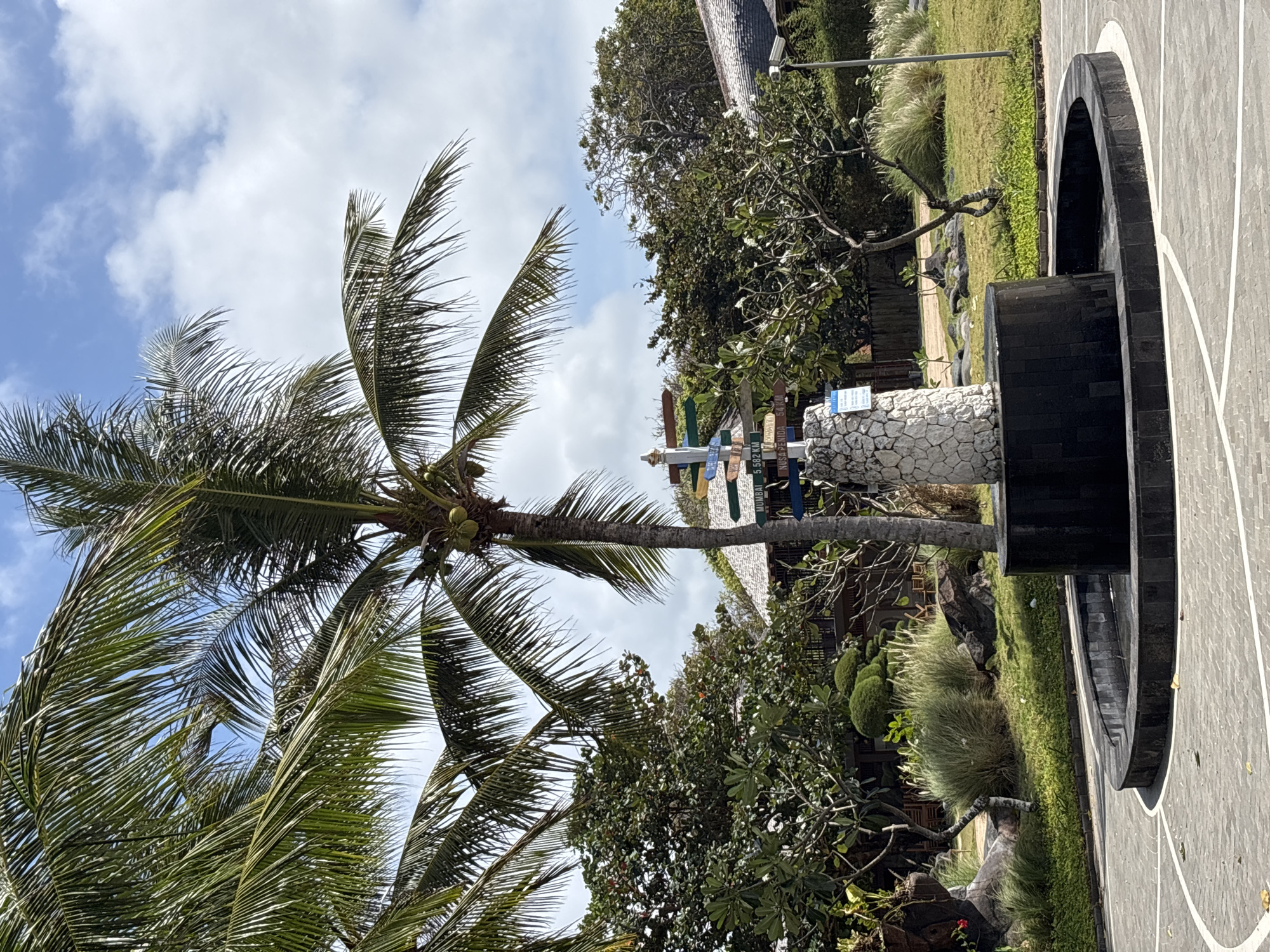 A directional signpost stands in front of a black, circular fountain in Kuta Selatan, Indonesia. The signpost, constructed with a stone base, displays multiple colorful signs pointing in various directions, some with distance markers. Palm trees with coconuts are visible in the foreground, with lush green foliage and trees in the background. Buildings with thatched roofs are also present. The sky is partly cloudy.