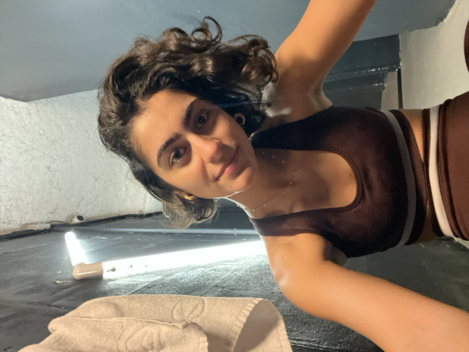 A young woman with curly brown hair smiles, looking slightly upward at the camera. She is wearing a brown athletic top with white stripes along the edges. A thin gold necklace is visible. She's indoors, the wall behind her is painted a textured white. A bright light and a white towel are visible in the foreground. The picture is taken at an angle, with the woman appearing to look down from above.