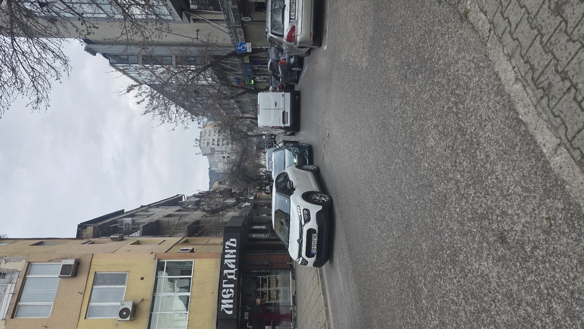 The image captures a street scene in Blagoevgrad, Bulgaria. The camera is positioned at street level, facing down the road. Parked cars line the right side of the street, including a white Citroen C4 Cactus with license plate "CB9247AE," a green car, and a white van. Further down the street are several other cars.
On the left side of the street, there is a yellow building with multiple windows and air conditioning units. The facade of the building features a business sign that reads "МЕРДАНЪ." Pedestrians are vaguely visible in the lower left corner.
The street itself is asphalt. The sky is overcast. Buildings are visible in the background. Leafless trees are present on both sides of the street.