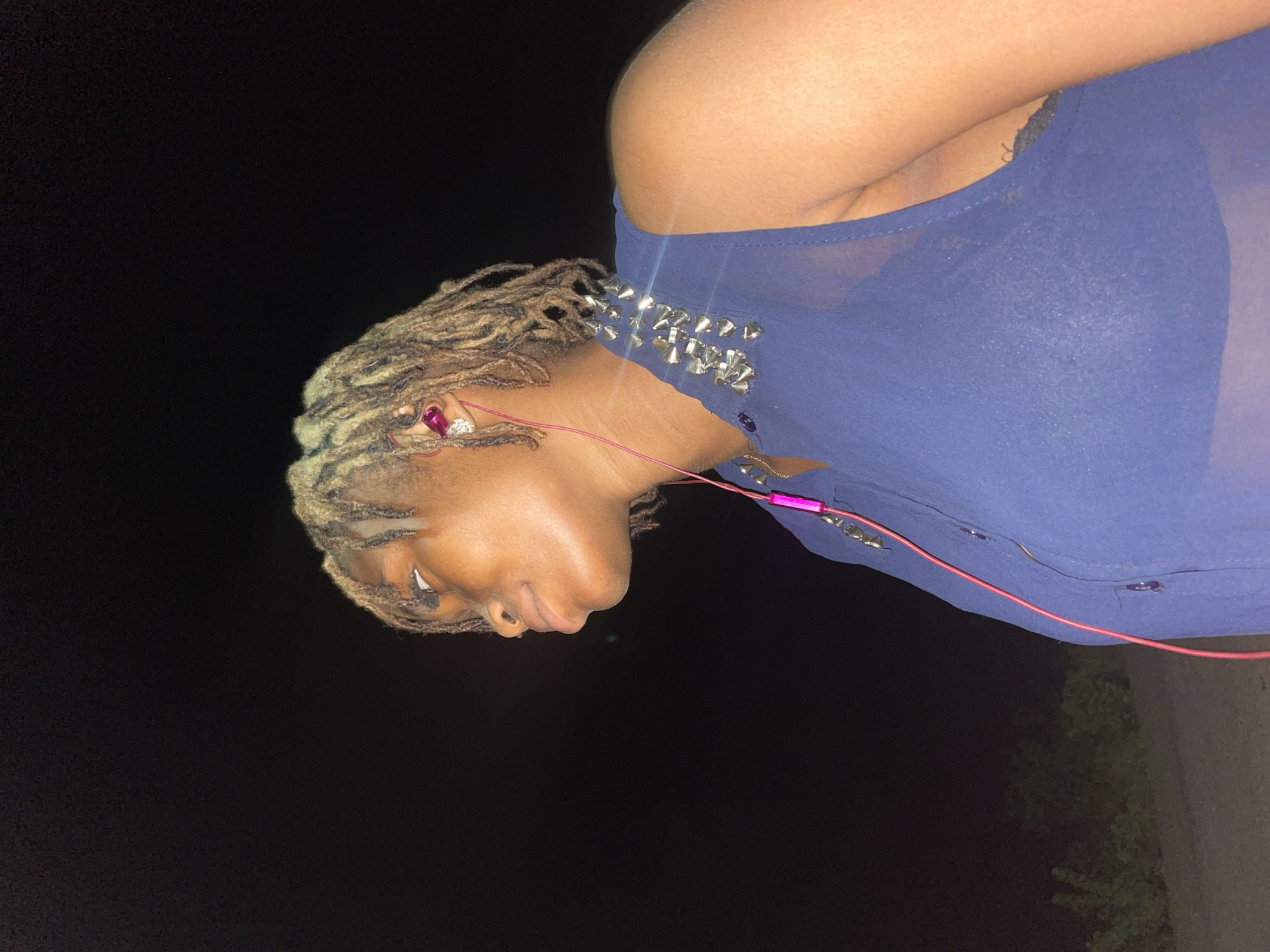 A person with blonde dreadlocks is wearing a blue top with silver spikes. The person has pink headphones in their ear. The background is dark. The image may have been taken in Ogbe, Nigeria.