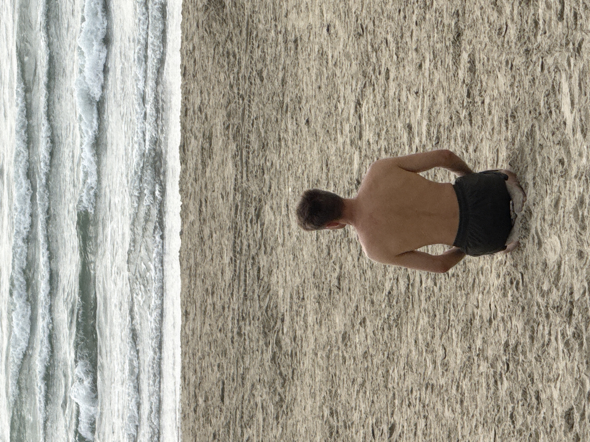 The image displays a shirtless adult male, viewed from the back, seated on a sandy beach. He wears dark-colored shorts and has short, brown hair. His knees are bent, and he faces directly towards the ocean. The foreground consists of textured, light brown sand. The background is dominated by the ocean, presenting multiple parallel lines of breaking waves with white foam and gray-green water. No other individuals or prominent objects are visible. The scene is located on a beach in Đà Nẵng, Vietnam.