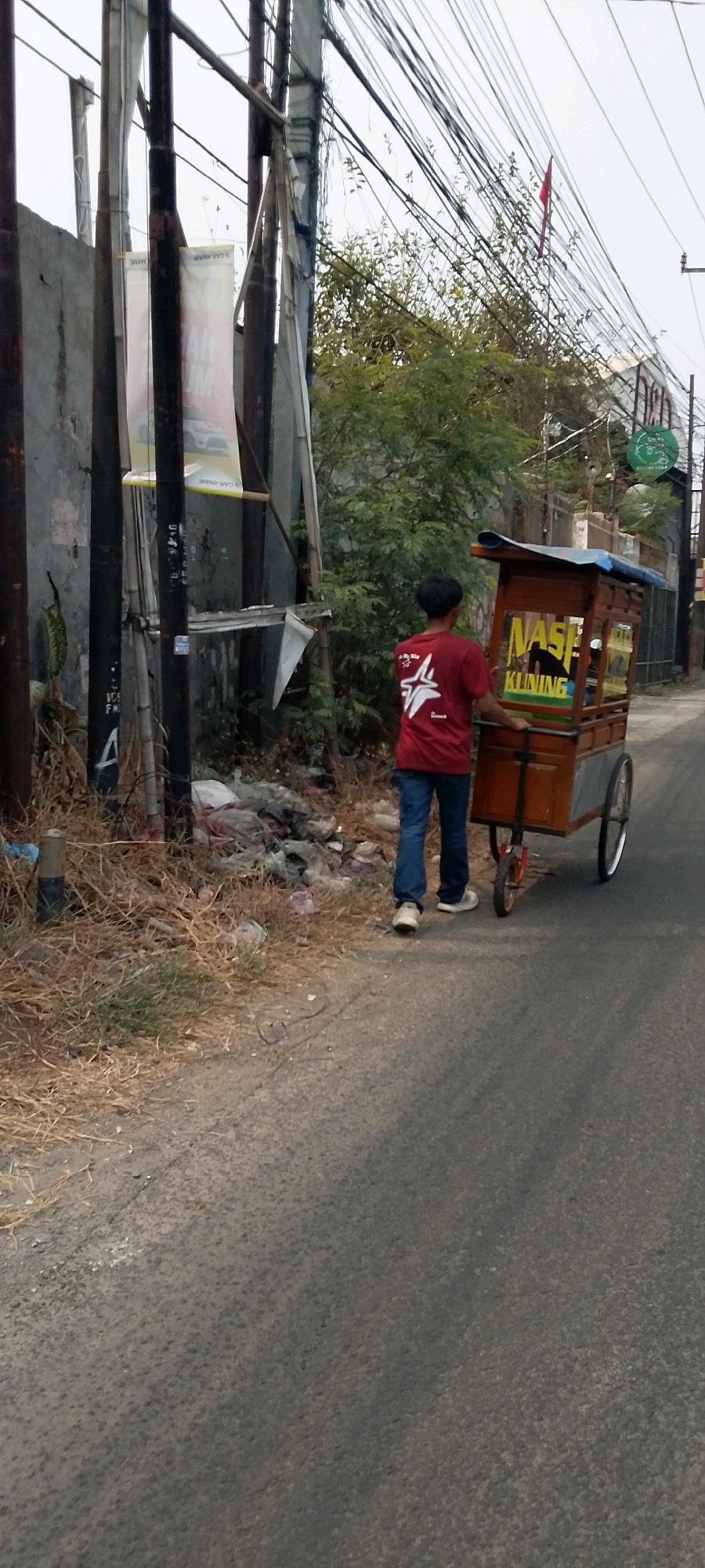 The image depicts a street scene in Bekasi, Indonesia. A young man wearing a red t-shirt and blue jeans is pushing a food cart. The cart is made of wood and has "Nasi Kuning" written on its side in yellow letters. In the background, there are trees, utility poles with many wires, a concrete wall, and a pile of trash. The road appears to be paved with asphalt. The overall impression is one of a typical urban street in Indonesia, with a mix of commercial activity and less-than-ideal environmental conditions.