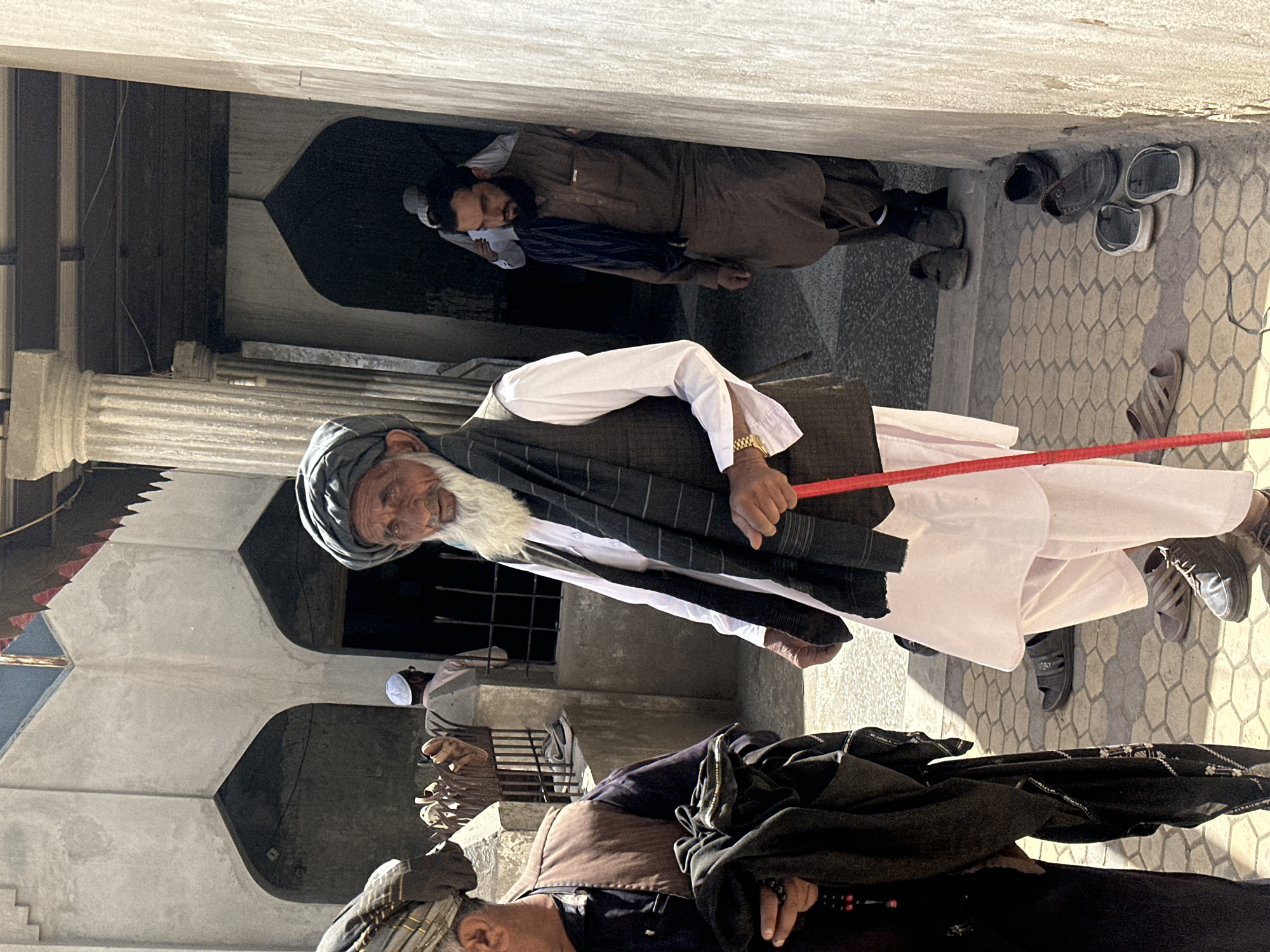 In the city of Kanozai, Pakistan, an outdoor scene unfolds. A group of men, primarily of Middle Eastern or South Asian descent, are present. The central figure, an elderly man with a long white beard and a dark turban, wears a white tunic, a dark vest, and a dark shawl. He holds a red-tipped cane and sports a gold wristwatch. To his left, another man, partially visible, wears a brown vest over a dark shirt and holds a folded black garment and a string of prayer beads. Further back, a man with a beard and a dark turban walks away from the camera, dressed in a brown tunic and trousers, with a dark scarf draped over his shoulders. Another individual in a white cap and shirt is seen in the background. The environment is characterized by concrete architecture, including arched doorways and fluted columns. The ground is paved with hexagonal tiles, and several pairs of sandals and shoes are scattered near a wall, suggesting a place of worship or public gathering. The lighting suggests it is daytime.