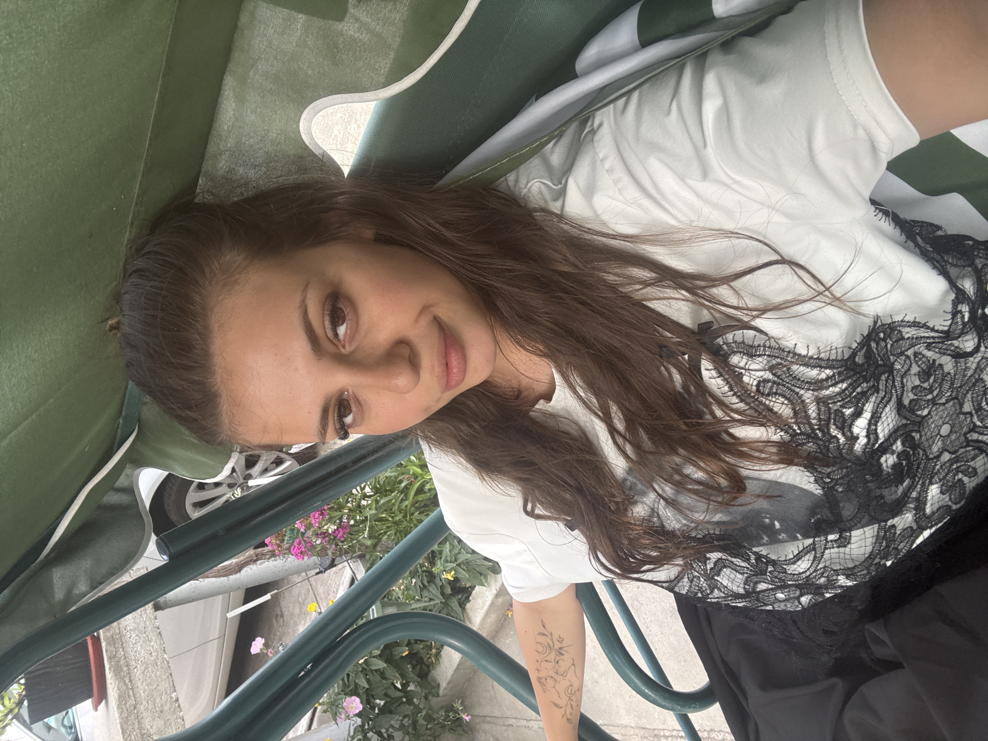 A young woman with long, dark brown hair pulled back into a loose bun is sitting on a green garden swing. She is wearing a white t-shirt with a black lace design on the front and a black skirt. She has a tattoo on her left arm. The swing is outdoors, with a blurred background showing pink and yellow flowers, a white car, and a potted plant. The overall setting suggests a residential outdoor area, possibly in Govedare, Bulgaria, given the information provided. The lighting is natural and appears to be daylight.