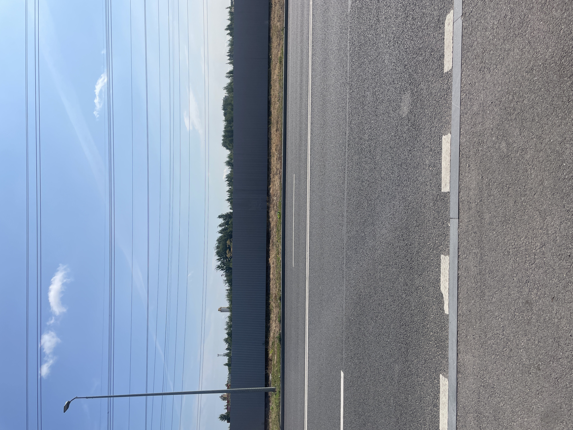 The image depicts an asphalt road with white lane markings under a clear blue sky. A tall streetlamp stands on the left side of the frame. In the background, a grey corrugated metal fence runs horizontally, bordered by a line of trees. Beyond the trees, industrial structures and what appear to be industrial chimneys are visible. Overhead, multiple power lines stretch across the sky, punctuated by a few wispy clouds. The scene is devoid of people or any overt activity, presenting a static, linear landscape. This appears to be a section of the road near the city of Pargolovo, Russia, likely a road leading to or from industrial areas.