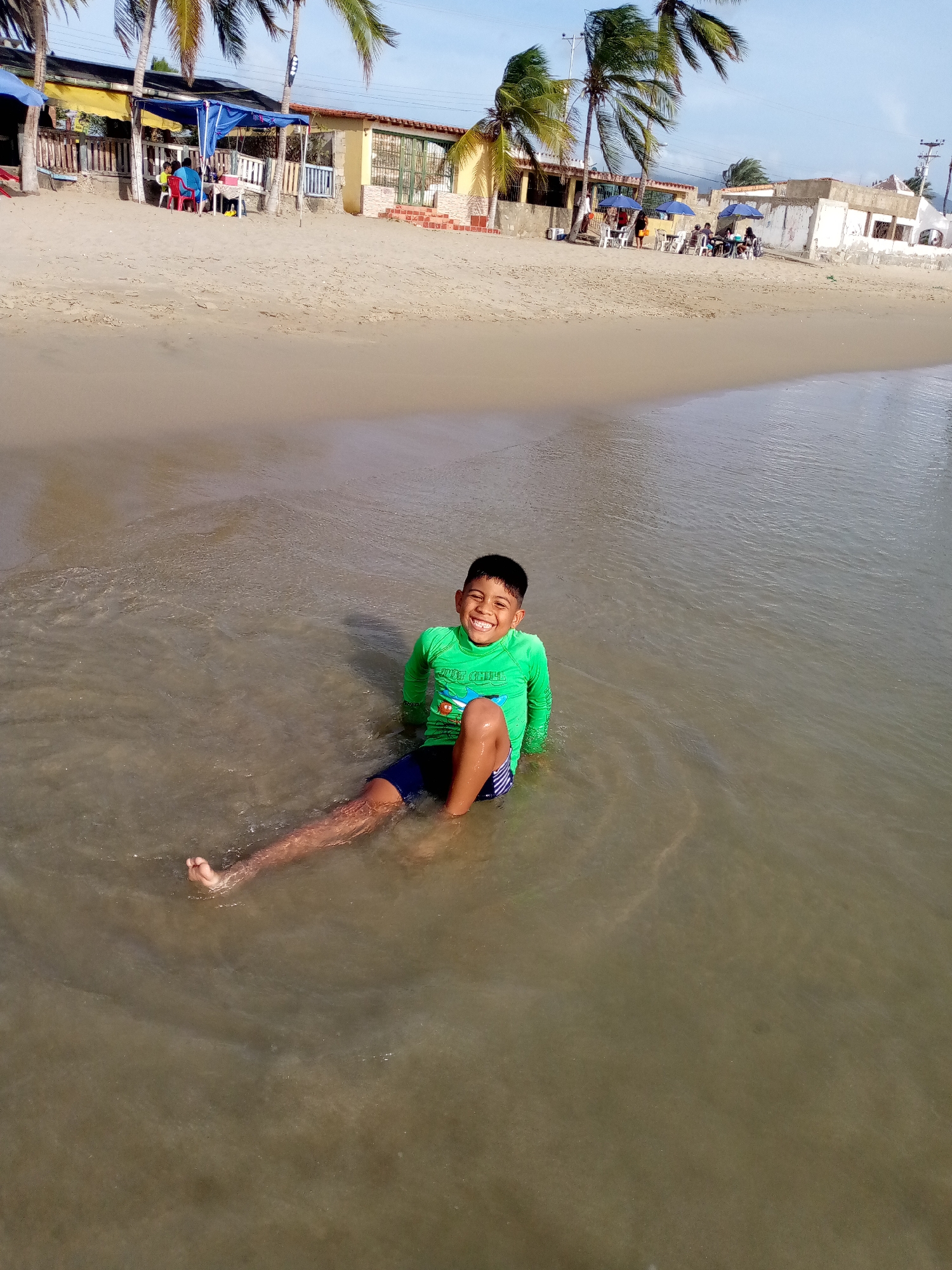 A vibrant scene unfolds on a sunny day at a beach in Juan Griego, Venezuela, featuring a young boy joyfully sitting in the shallow, calm water. He is positioned in the foreground, facing the camera with a wide, infectious smile, conveying pure happiness. He wears a bright green long-sleeved rash guard, upon which the text "JUST CHILL" is partially visible on his chest, and blue swim trunks. His legs are stretched out into the clear water, which gently ripples around him, revealing the sandy bottom beneath.
Behind the boy, the expansive sandy beach stretches towards a line of buildings and palm trees. On the left side of the midground, several people are seated under a blue and yellow canopy, likely enjoying a meal or refreshments at tables and chairs. Further along the beach to the right, more groups of individuals are visible, relaxing under blue beach umbrellas and additional canopies, also seated at tables with white plastic chairs. The background reveals a series of low-rise beachfront buildings, some with yellow or white facades, and architectural details like red brick bases and green window frames. Several tall palm trees are scattered along the shore, their fronds adding to the tropical ambiance. The sky above is bright and clear, with only a few wispy clouds, indicating warm and pleasant weather. The overall scene depicts a lively and relaxed day by the sea.