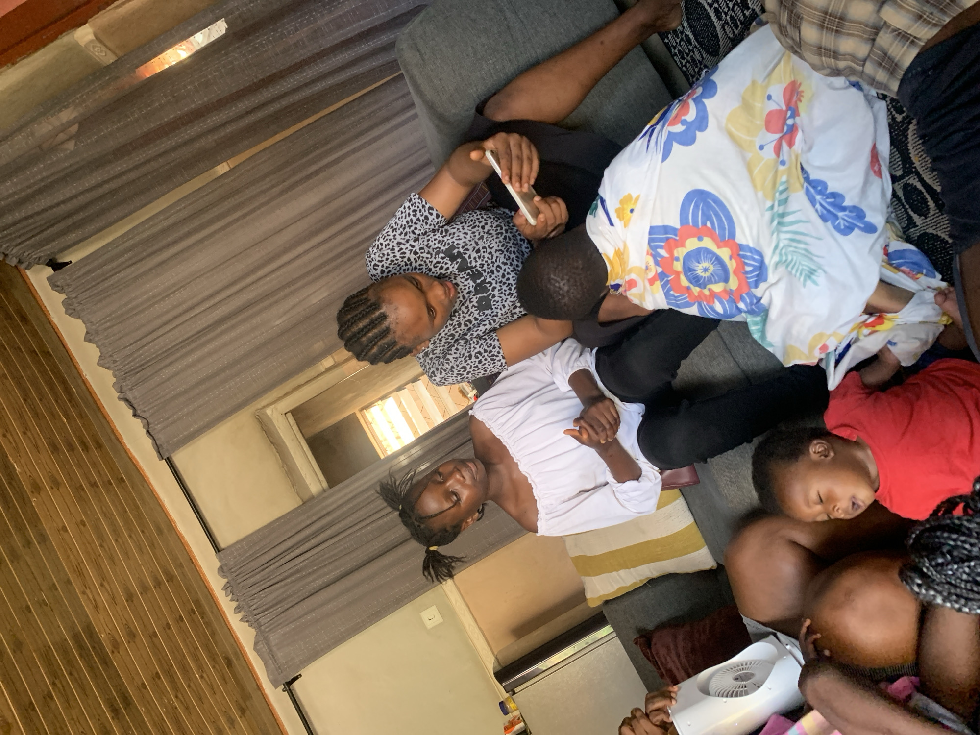 In Sabon Tasha, Nigeria, a group of people are gathered in a living room with a wooden slatted ceiling. On a gray couch, two women are seated. One woman, on the left, wears an off-the-shoulder white blouse and has her hair in two pigtails. She is looking down and appears to be talking. The other woman, on the right, has braided hair and wears a black and white leopard-print t-shirt. She is holding a smartphone in her hands and looking at it, possibly showing something to a young child seated in front of her. This child is wrapped in a white blanket with a colorful floral pattern. To the left of the woman in white, a baby wearing a red t-shirt is lying down, with its eyes closed and mouth open. A small white fan is placed on the couch to the left of the baby. The room also features gray curtains and a refrigerator to the left of the couch.