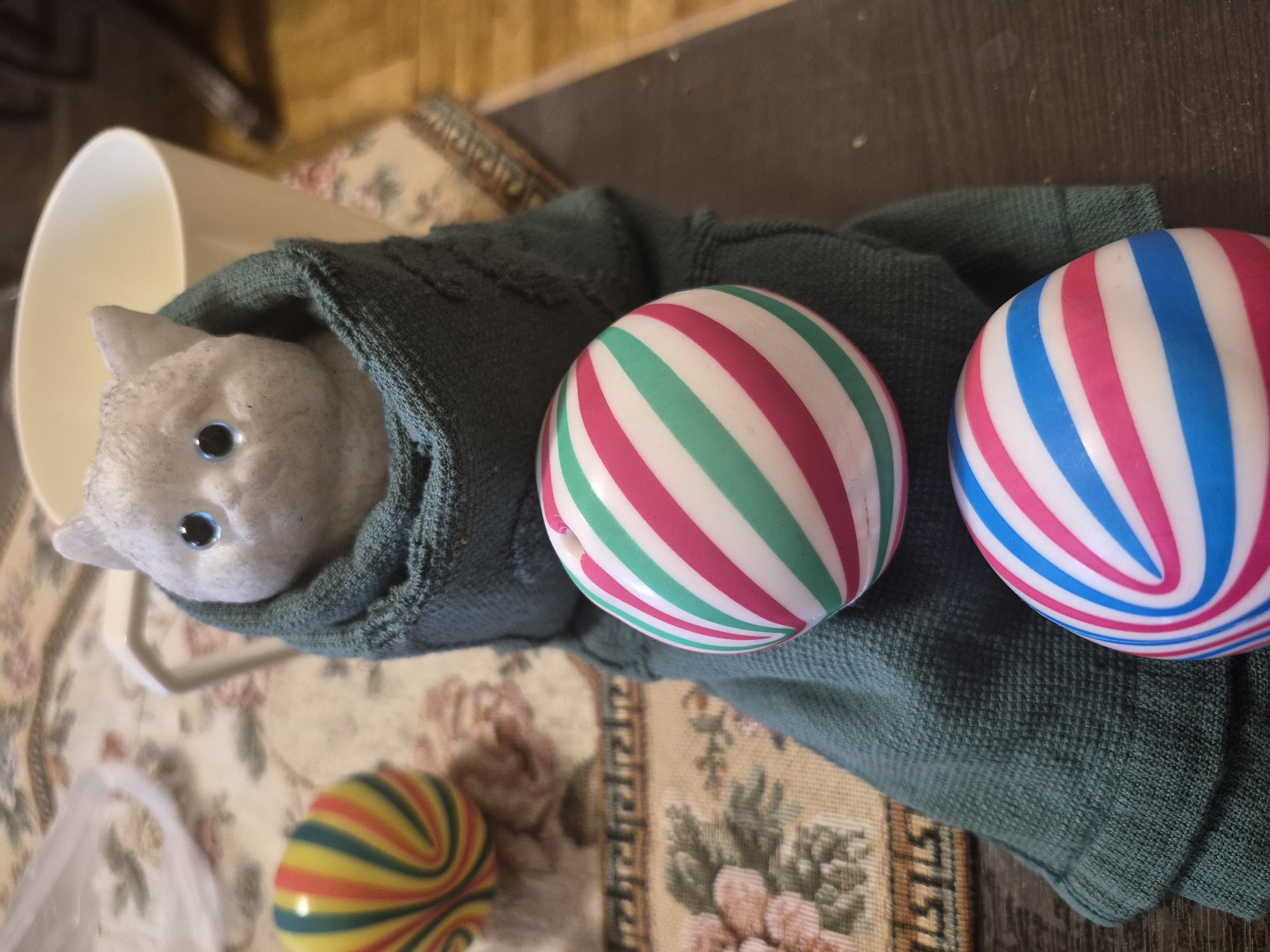 The image depicts a still life arrangement on a dark wooden surface, with a patterned tablecloth in the background. The primary subjects are a small grey cat figurine and three striped balls. The cat figurine, with blue eyes, is partially wrapped in a green knitted garment that resembles a sweater or scarf. In front of the cat are two striped balls, one with pink and green stripes, and another below it with pink and blue stripes. To the left of the cat and slightly further back is a third ball, with multicolored stripes of yellow, red, green, and blue. In the background, a white cup or vase is visible, along with portions of the patterned tablecloth and a wooden floor. The scene is lit with indoor lighting. There is no information available regarding the location being in Yerevan, Armenia, as the image context does not provide any such details.