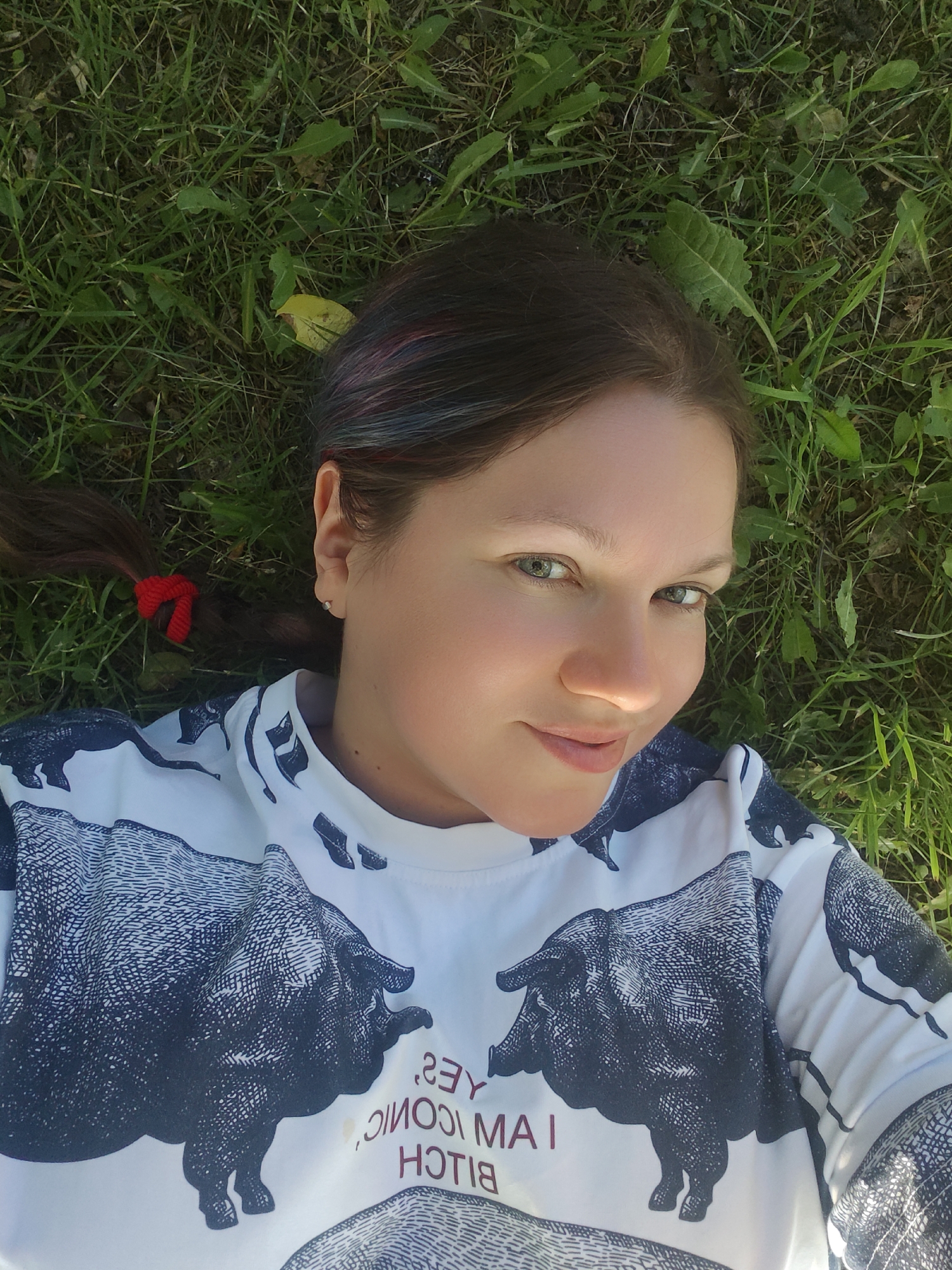 An adult female is depicted lying on her back on a surface of green grass in Dubna, Russia, gazing directly at the camera. She has a fair complexion, dark hair with subtle colorful highlights, and green eyes, displaying a slight, direct smile. Her hair, styled in a ponytail to her left, is secured with a red hair tie. A small silver stud earring is visible in her left earlobe. She wears a white t-shirt adorned with a large, black, etched-style graphic of a boar. The t-shirt also features reversed red text, which reads "YES, I AM ICONIC, BITCH". The immediate environment consists of green grass interspersed with various broadleaf plants and one yellowed leaf. The scene is illuminated by natural daylight.