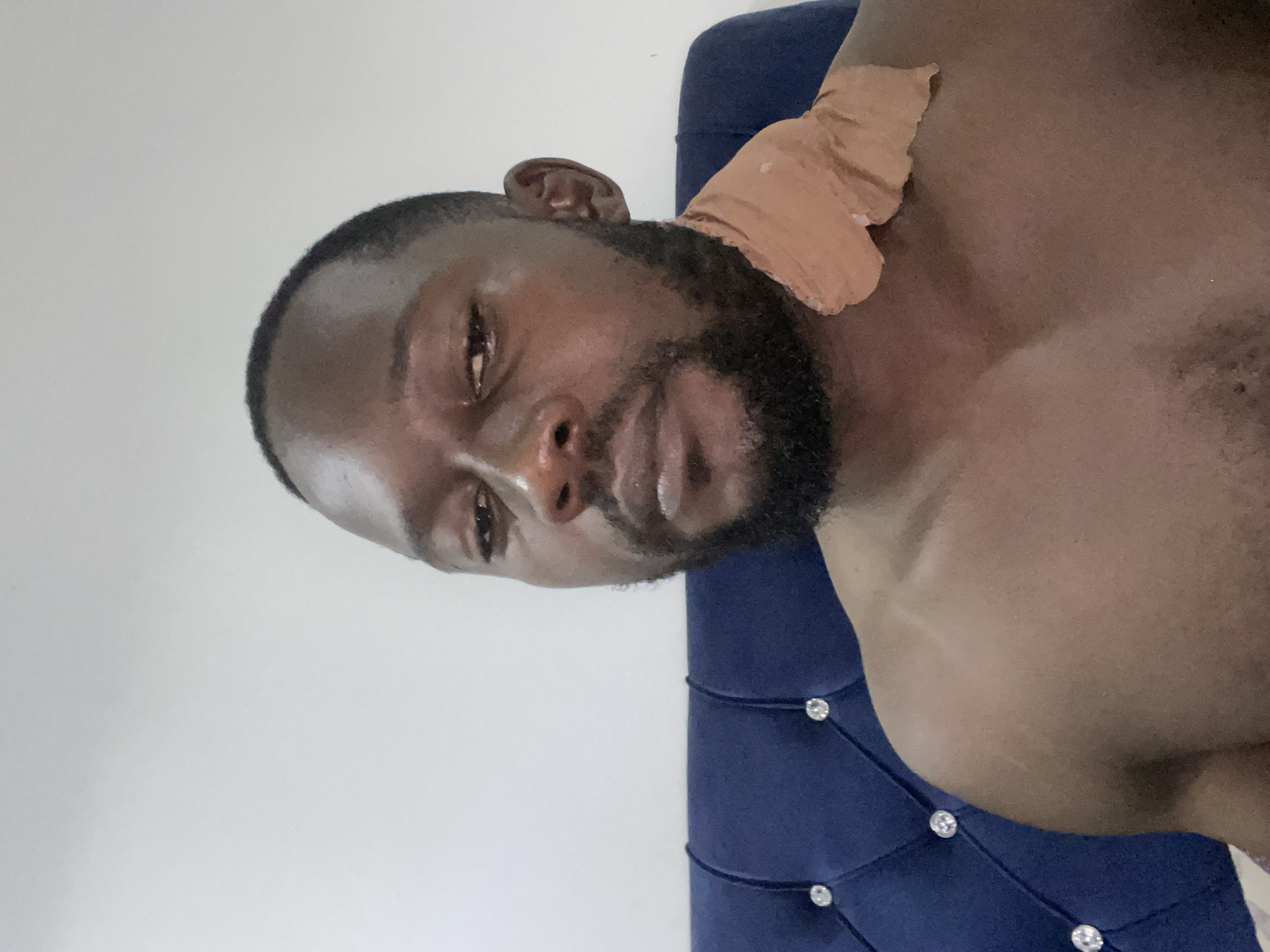 A man with dark skin, a beard, and short black hair is pictured from the chest up, looking directly at the camera with a neutral expression. He is shirtless, and a tan bandage is visible on his upper chest and neck area, extending over his right shoulder. The background consists of a plain white wall and a portion of a dark blue, tufted headboard with crystal-like embellishments. The lighting appears to be artificial and casts soft shadows. There is no indication of the specific location within Abuja, Nigeria.