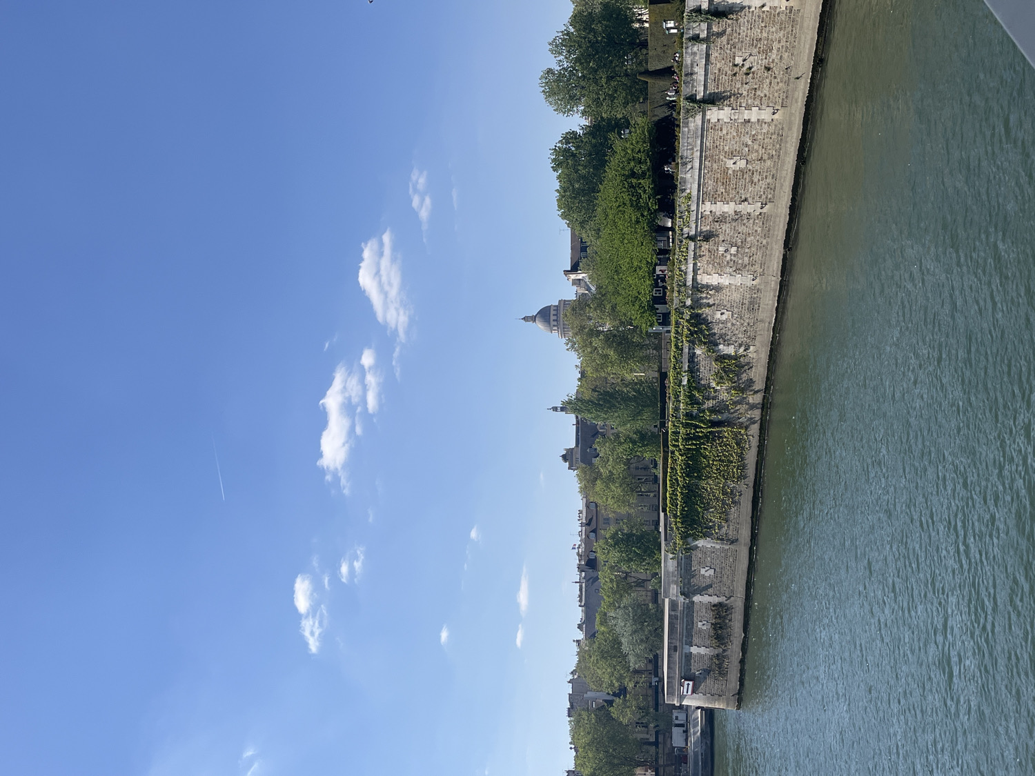 This image captures a bright, sunlit view along a wide river, likely the Seine in Paris, France. The river occupies the right side of the frame, presenting a calm, greenish-blue surface. On the left, a robust stone embankment lines the riverbank, featuring multiple tiers and partially adorned with lush green vegetation, possibly ivy or vines, clinging to its walls.
Above the embankment, a dense array of mature green trees forms a prominent feature, behind and through which various buildings are visible. A distinct architectural element, a large dome-shaped structure, stands out against the sky, strongly suggesting a significant Parisian landmark such as Les Invalides. Further along the riverbank, other characteristic Parisian buildings with their grey roofs and classical facades can be seen.
The sky above is a vibrant, clear blue, dotted with scattered, fluffy white cumulus clouds. A faint, white airplane contrail stretches diagonally across the upper left portion of the sky. The scene depicts a serene, bright day, likely morning or early afternoon, with ample sunlight illuminating the cityscape and river. There are no specific activities or discernible text visible in the frame. The architectural cues and the provided context firmly place the location in Paris, France.
