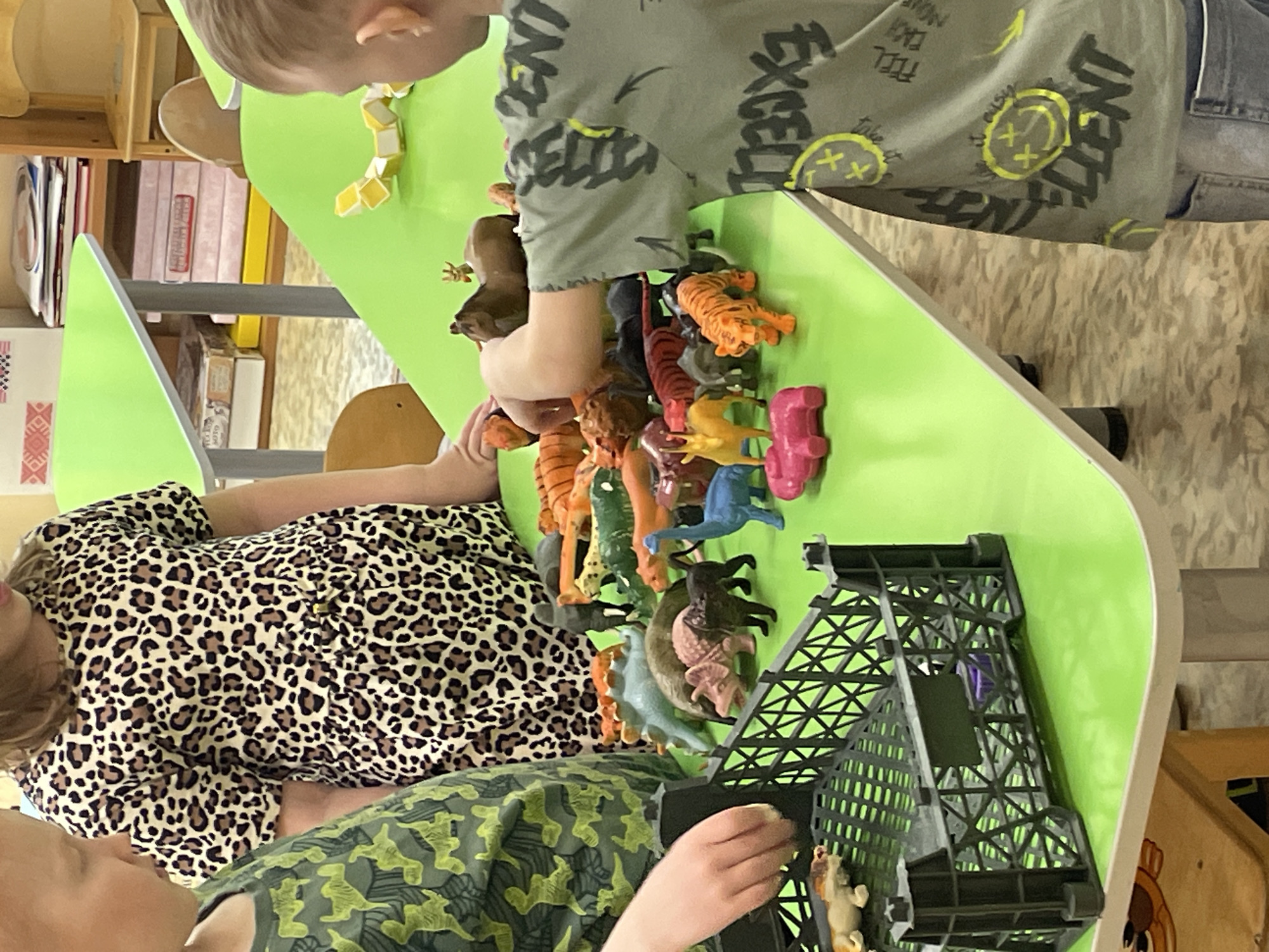 Two children play with toy animals on a green table. One child wears a leopard-print shirt, and the other wears a patterned green shirt. The toys include various animals like tigers, dinosaurs, and a lion. A black cage-like structure is also present on the table. The setting appears to be indoors, possibly a classroom or playroom. The location is not identifiable as Izhevsk, Russia, from the given image.