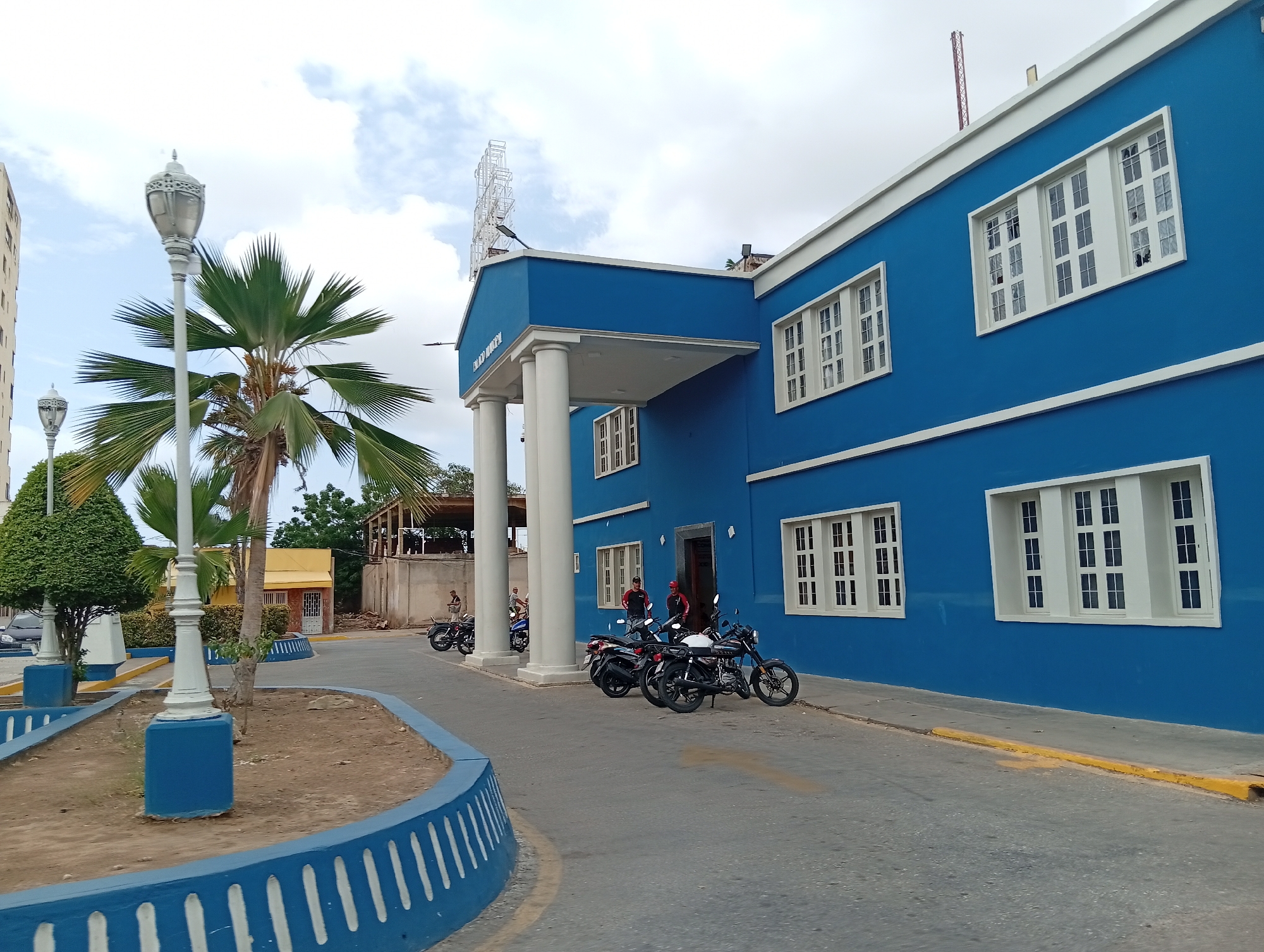 The image shows a bright blue building with white trim and windows, located in Juan Griego, Venezuela. The building has a covered entrance supported by white columns. A metal lattice structure is mounted on the roof above the entrance. Several motorcycles are parked in front of the building. Two people stand near the entrance of the building. A palm tree and a white street lamp are in the foreground on the left. A curb with blue and white stripes borders a landscaped area in front of the building. The sky is partly cloudy.