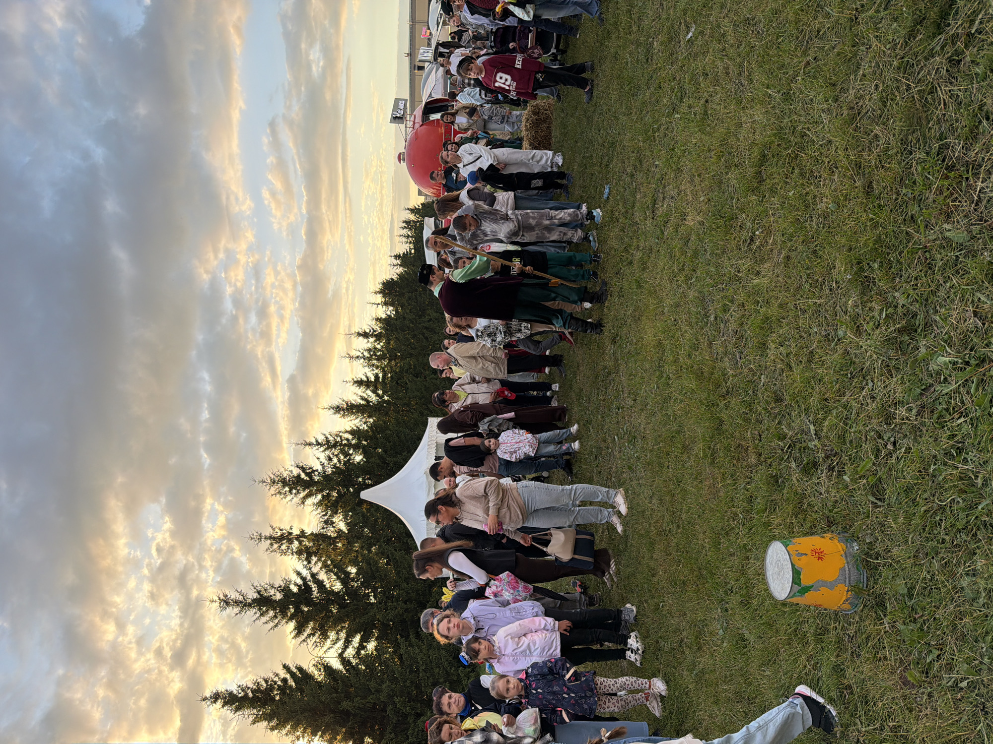 In an outdoor grassy field, a crowd of people, including many children, stand in clusters. The background features a line of evergreen trees and a few tents and food trucks, one of which is bright red and dome-shaped. The sky above is filled with dramatic, cloudy skies illuminated by the golden hues of sunset. In the foreground, a person in jeans and sneakers is walking away from the camera, and a worn, yellow and orange bucket sits on the grass.