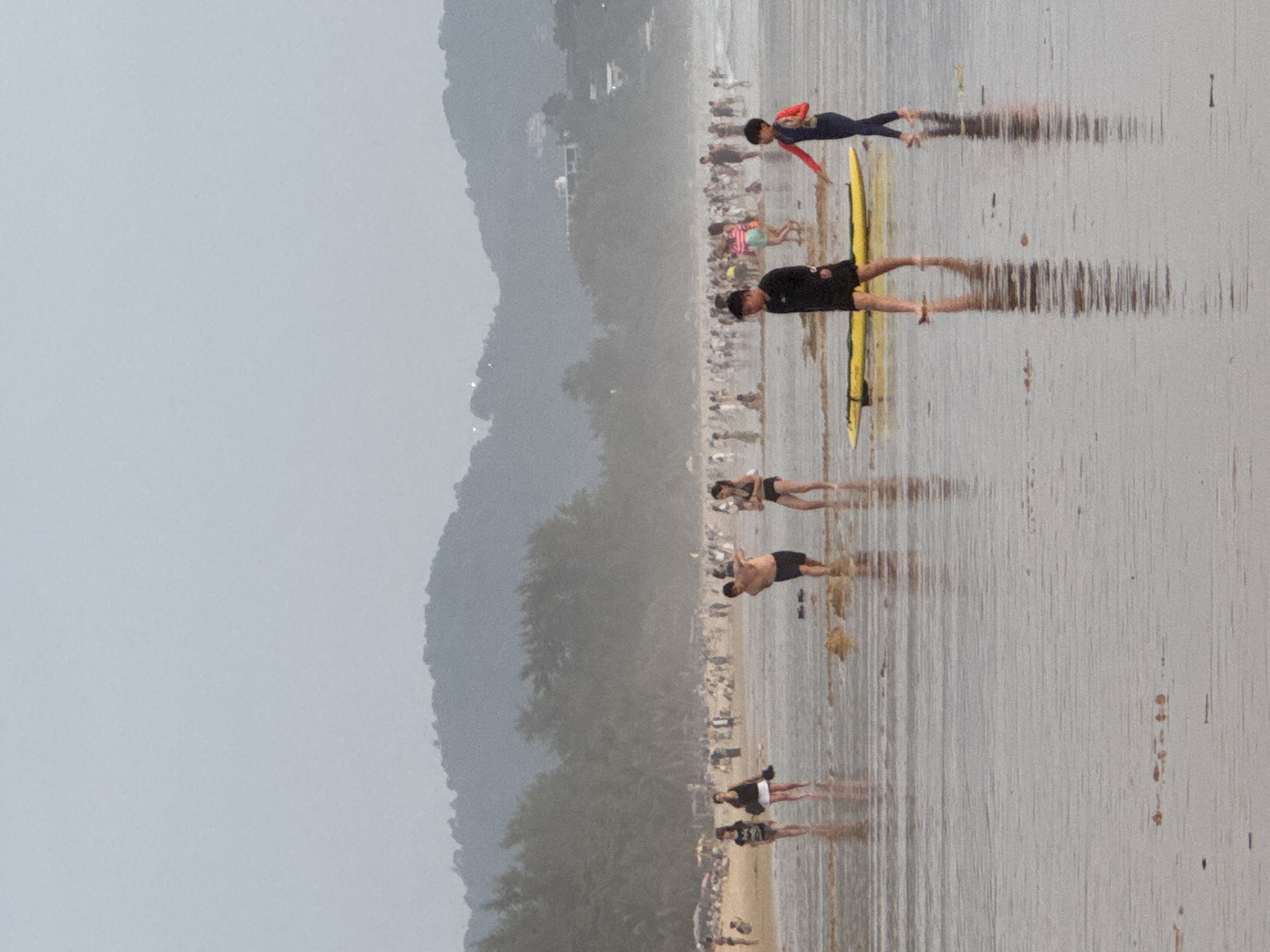 The image depicts a crowded beach scene on a hazy day. In the foreground, several individuals are walking on the wet sand and shallow water. A boy in a dark shirt and shorts walks towards the left, with a yellow surfboard lying on the sand to his right. To his right, a child in a blue and red wetsuit walks towards the right, with their arm outstretched. Further in the foreground, a man in dark shorts is wading in the water, facing away from the camera. To his left, two women walk on the wet sand, one in a black and white striped top and skirt, and the other in a black top and white skirt. Behind them, a dense crowd of people are visible on the beach, with many appearing to be sitting or standing under umbrellas. The background shows a treeline, followed by rolling hills or mountains, possibly with some buildings visible through the haze on the upper slopes. The overall impression is of a busy recreational area on a cloudy or humid day.