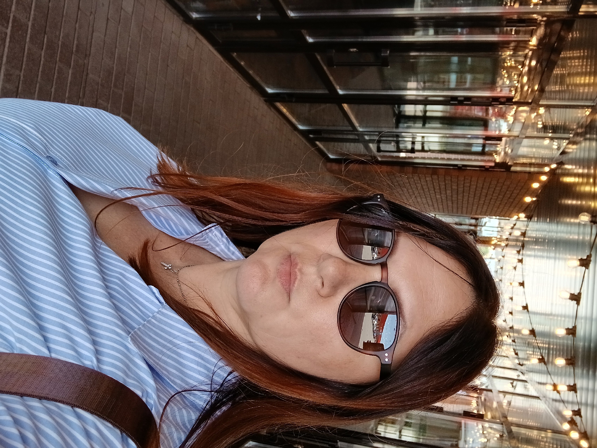 A medium shot captured from a low angle shows a woman standing outdoors in what appears to be a covered walkway or an arcade. The woman, facing the camera, has long brown hair and is wearing sunglasses, a light blue and white striped button-down shirt, and a silver cross necklace. Her right shoulder strap of a brown bag is visible across her shirt. The background consists of a brick wall on the right and a series of large glass doors and windows on the left, reflecting a row of parked cars and buildings. String lights are visible hanging overhead, illuminating the walkway. The ground is paved with what looks like brick or cobblestone. The overall lighting suggests it is daytime, with the string lights adding a decorative element.