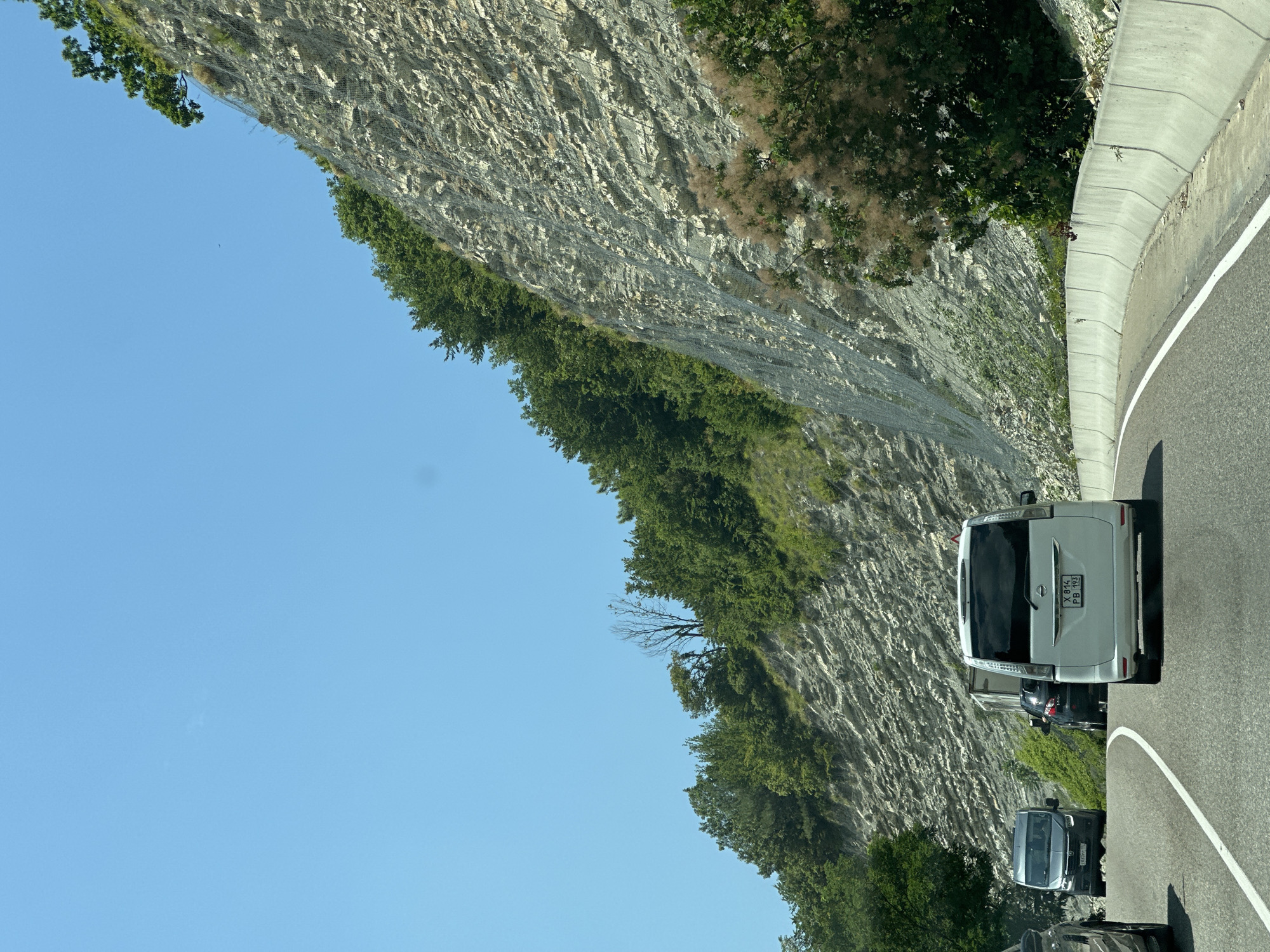 The image shows a scenic view of a road curving alongside a steep, rocky mountainside covered in lush green trees. A clear blue sky stretches above. Several vehicles are on the road, suggesting a travel route. In the foreground, a white minivan with license plate "X 814 PB 193" is traveling forward. Behind it, a dark vehicle and a grey minivan are also visible. The mountain slope is protected by a wire mesh to prevent rockfalls. The presence of this road and its unique geography suggests it could be a popular route for tourism or transportation in a mountainous region like Kroyanskoye, Russia, known for its natural beauty and dramatic landscapes.