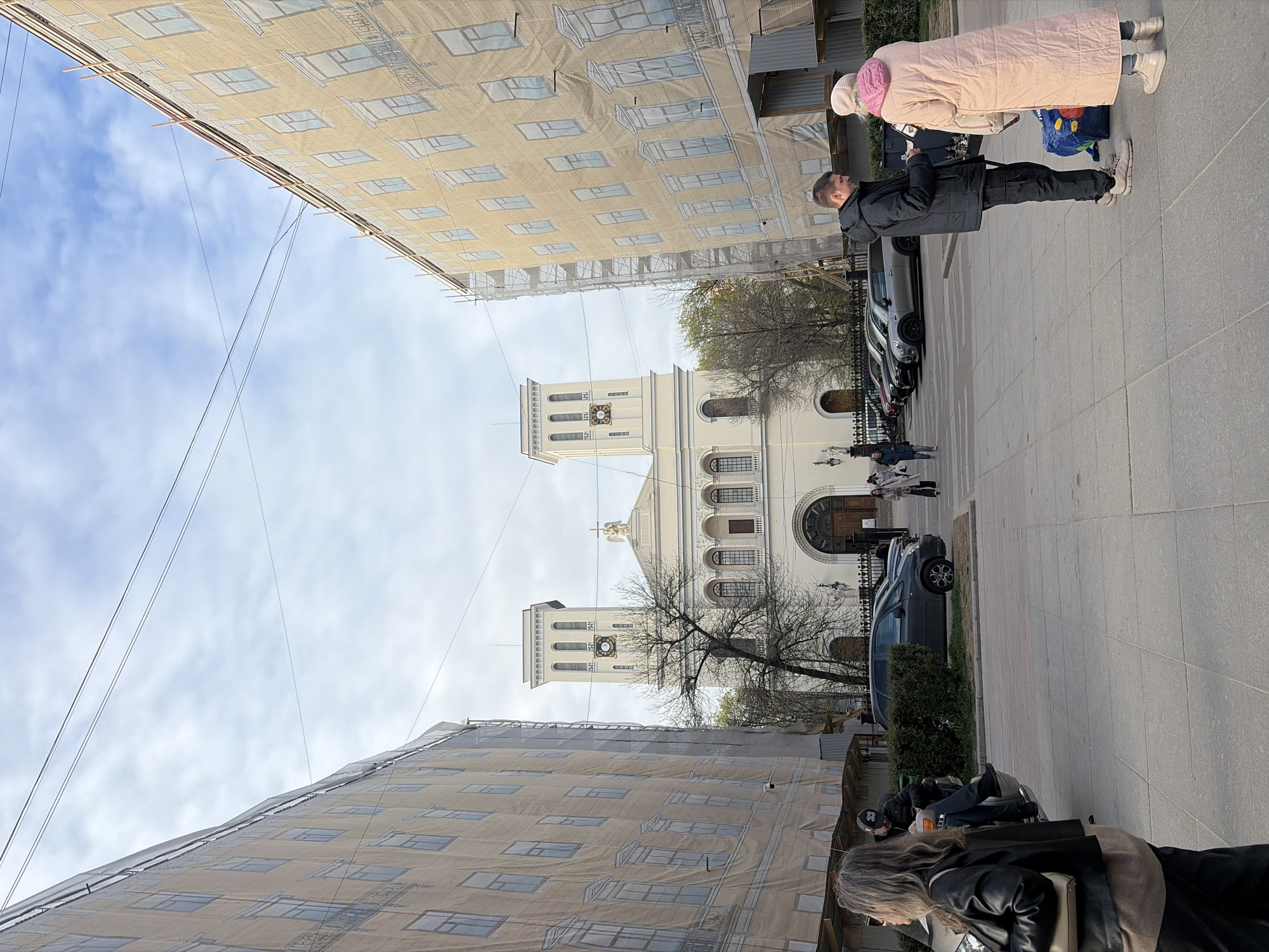 The image captures a street scene in Saint Petersburg, Russia. The focal point is the Chesme Church, a two-towered structure with a cream-colored facade and arched windows. Flanking the church on either side are buildings covered in scaffolding and protective scrims, printed with a graphic of the building facades.
On the right side of the frame, a sidewalk hosts pedestrian activity, including a man in a black jacket talking to a woman in a pink coat, as well as other pedestrians. Several cars are parked along the street, including a dark-colored sedan. A motorcycle and rider are visible in the foreground on the right. The sky is partly cloudy.
