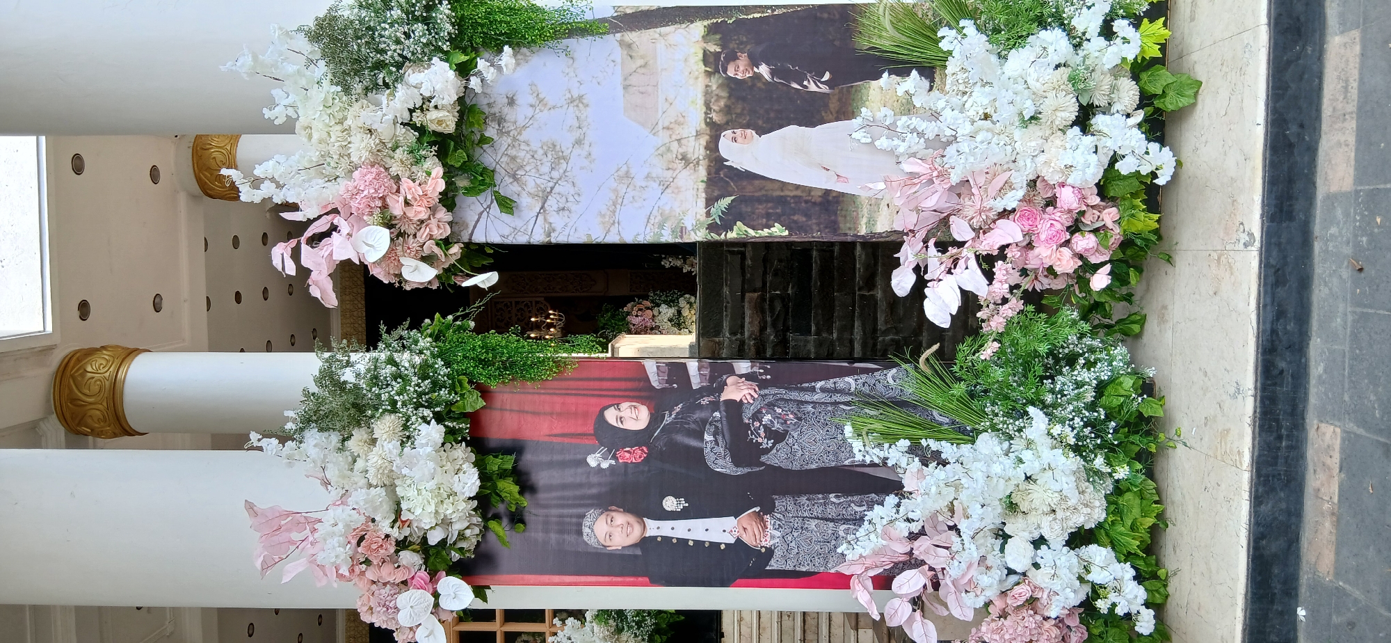 The image shows a wedding decoration setup outside a building, likely a venue in Samarinda, Indonesia. Two large wedding portraits are displayed prominently. The portrait on the left features a couple dressed in traditional attire. The man wears a black formal jacket over a white shirt and batik pants, with a patterned peci on his head. The woman is in a black hijab and a matching patterned outfit, adorned with jewelry. The portrait on the right shows a bride in a white wedding gown and veil, standing beside a groom in a black suit.
The portraits are flanked by elaborate floral arrangements, predominantly in white and blush pink hues, with abundant greenery. These arrangements are positioned in front of large white pillars, one of which has a decorative gold band near the top. Behind the portraits, a set of stone stairs leads up to an entrance, suggesting this is the entryway to the event space. The overall setting appears to be an outdoor public space, possibly a plaza or courtyard adjacent to the building, with cobblestone or tiled flooring visible at the bottom of the frame. The lighting suggests it is daytime.