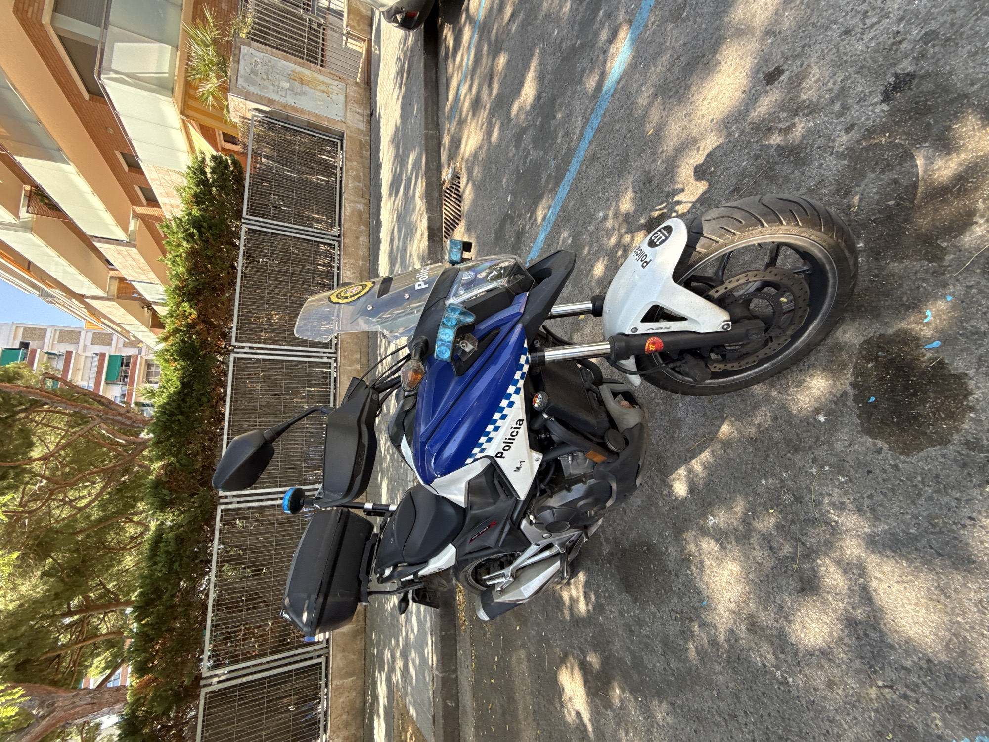 The image shows a police motorcycle parked on a paved surface, possibly a street or parking lot. The motorcycle is predominantly white and blue, with "Policia" written on the side. It has saddlebags on the rear and a windshield at the front. The scene appears to be outdoors during the daytime, with shadows cast on the ground, indicating sunlight. The background includes a building with balconies and trees. The ground has some blue markings, possibly indicating a parking space. The overall setting suggests an urban environment in a location with a Mediterranean climate, given the trees and building style, consistent with Blanes, Spain. No people are visible in the image. The time of day appears to be late afternoon, judging by the length and angle of the shadows.