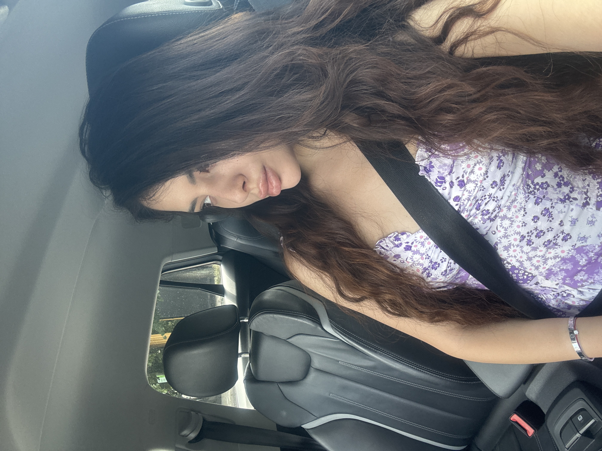 A young woman with long, wavy dark hair is seated in the passenger seat of a car. She is wearing a purple and white floral print top and a seatbelt. Her right arm, adorned with a silver bracelet, rests on the car's seat. The car's interior is visible, including the leather seats and window. The background visible through the window is out of focus but shows signs of greenery and possibly a building. There is no indication the image was taken in Plovdiv, Bulgaria.