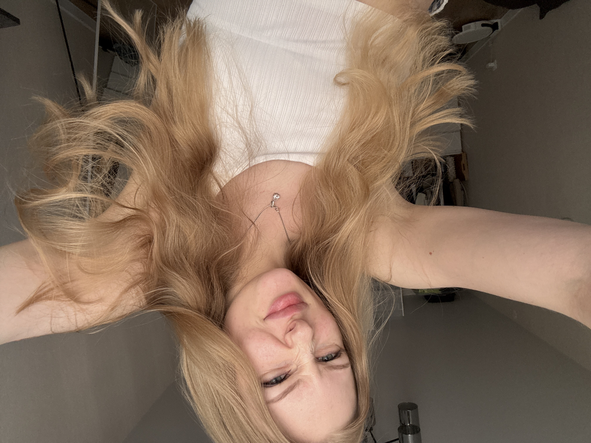 A selfie of a woman with long blonde hair, wearing a white top and a necklace, is taken from an upside-down perspective. The location is not identifiable as Perm, Russia based on the available visual information. The woman is squinting her eyes and pursing her lips. The background is a neutral color.