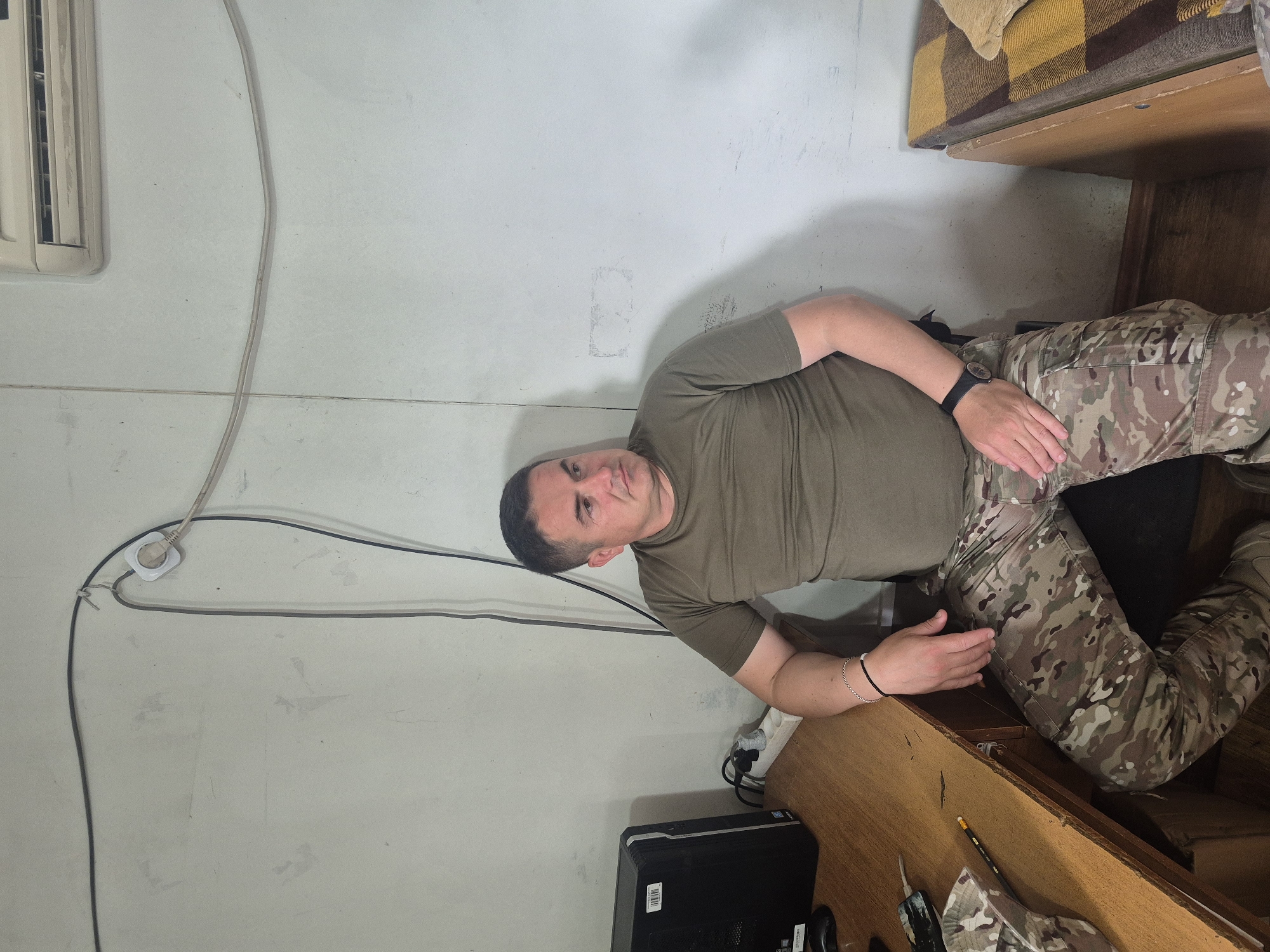A man wearing a green t-shirt and camouflage pants sits in what appears to be an indoor setting, possibly a storage room or office. The room has white walls with electrical wiring visible. There is a desk or shelf with items such as a computer tower, writing utensils, and other objects. A bunk bed or similar structure is present above the desk area. The man is wearing a watch on his left wrist and a bracelet on his right. The exact location within the city of Tbilisi, Georgia cannot be determined from the image alone.