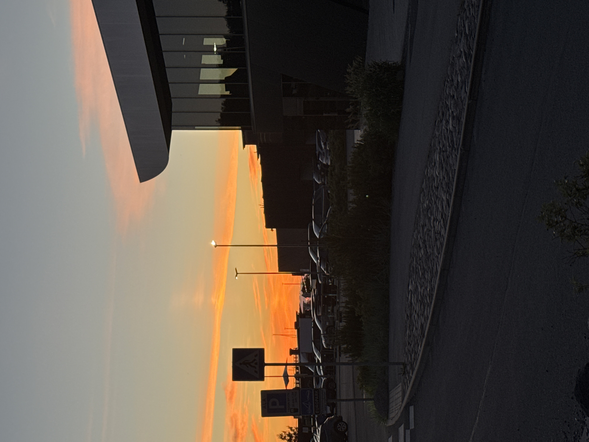 The image depicts an outdoor parking lot and the exterior of a modern building at sunset in Tallinn, Estonia. The sky is a vibrant mix of orange and pink hues, with wispy clouds. In the foreground, a paved road curves to the left, bordered by a cobblestone pathway and a strip of greenery. Several cars are parked in the lot, some silhouetted against the bright sky. Two signs are visible near the parking lot, one with a "P" indicating parking and another showing a pedestrian crossing symbol. A street lamp is visible on the right side of the image, illuminating a small area of the parking lot. The building on the right has large glass windows reflecting the sunset and the surrounding environment. The overall impression is one of a serene, urban evening scene.