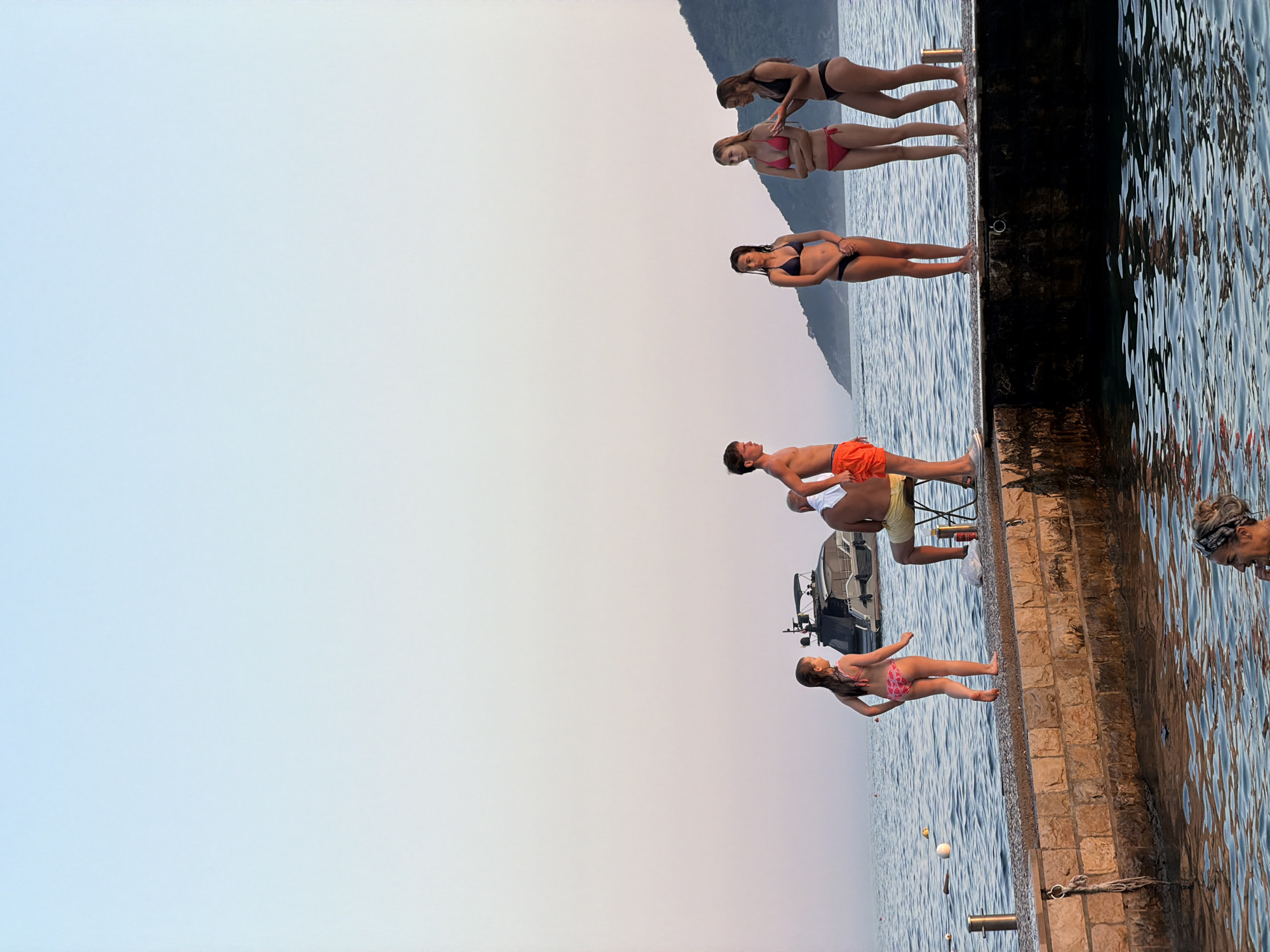 This media file depicts a scene at Boreti, Montenegro. Several people, primarily young women and a few individuals who appear to be men and children, are positioned along a stone pier and on the water.
The subjects are dressed in swimwear, suggesting a recreational activity at a seaside location. Some individuals are standing on the pier, while others are interacting with the water. One young woman in a dark bikini is standing with her hands on her hips, looking out at the water. Further up the pier, two young women are embracing, with one in a black bikini and the other in a pink bikini. Below them, another young woman in a dark bikini stands with her arms crossed. A young man in orange shorts is partially visible, and a child in a pink and white patterned bikini is walking along the pier towards the water. A woman with her hair tied back is visible near the edge of the pier.
The setting is a waterfront area with a calm body of water, likely the Adriatic Sea, given the location context. A boat is docked in the water, and the background shows a hazy mountainous landscape. The time of day appears to be late afternoon or early evening, indicated by the soft, diffused light and the warm tones of the sky. The weather seems pleasant and clear. The overall mood conveyed is one of leisure and enjoyment of a summer day at the beach. There is no visible text in the image.
