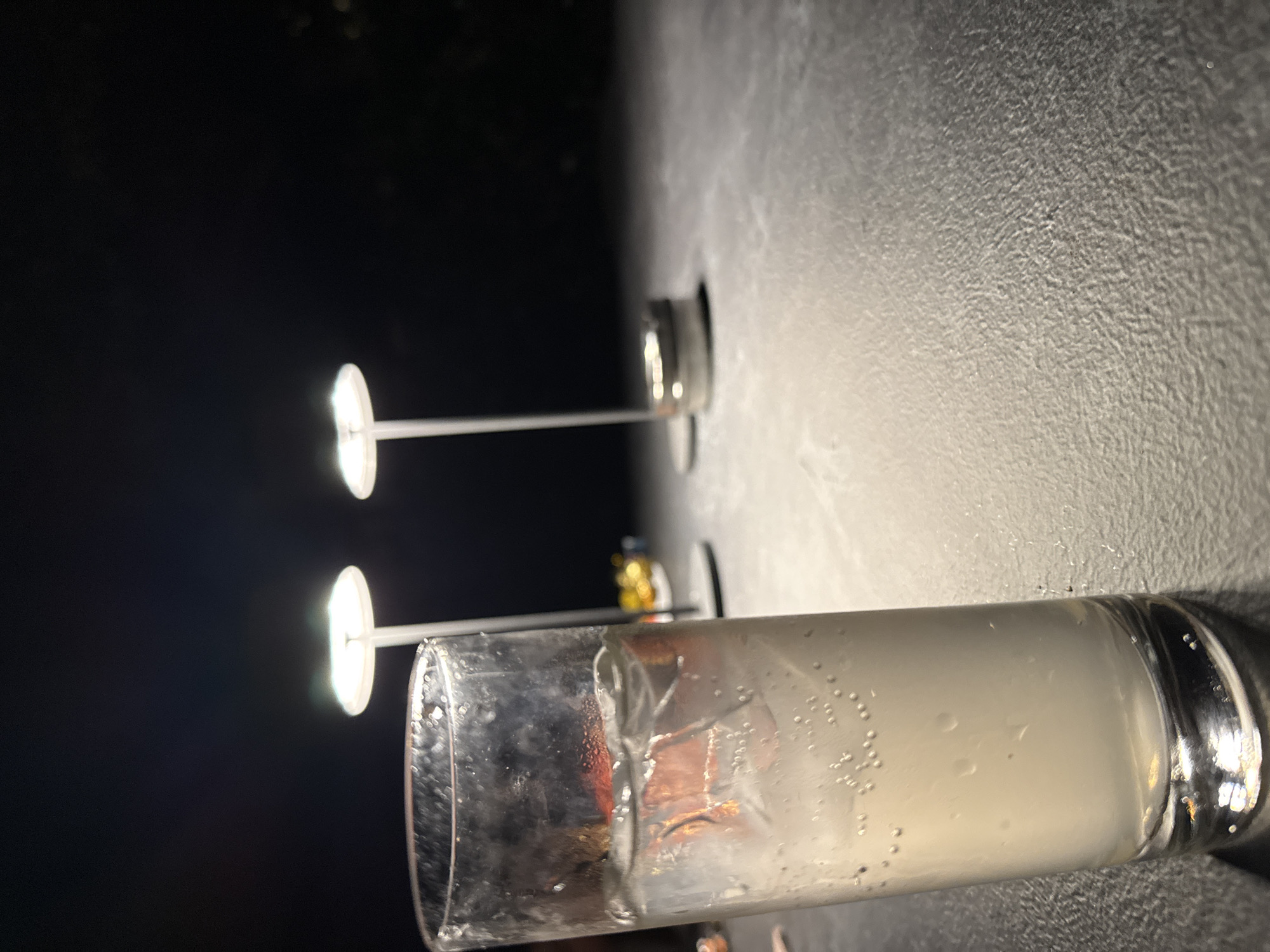 The image shows a glass of liquid with ice sitting on a textured surface. The liquid is milky and contains many small bubbles. Reflected in the glass are two street lights, suggesting the scene is located outdoors at night. The location is unknown but inferred as the city of Castel Gandolfo, Italy.