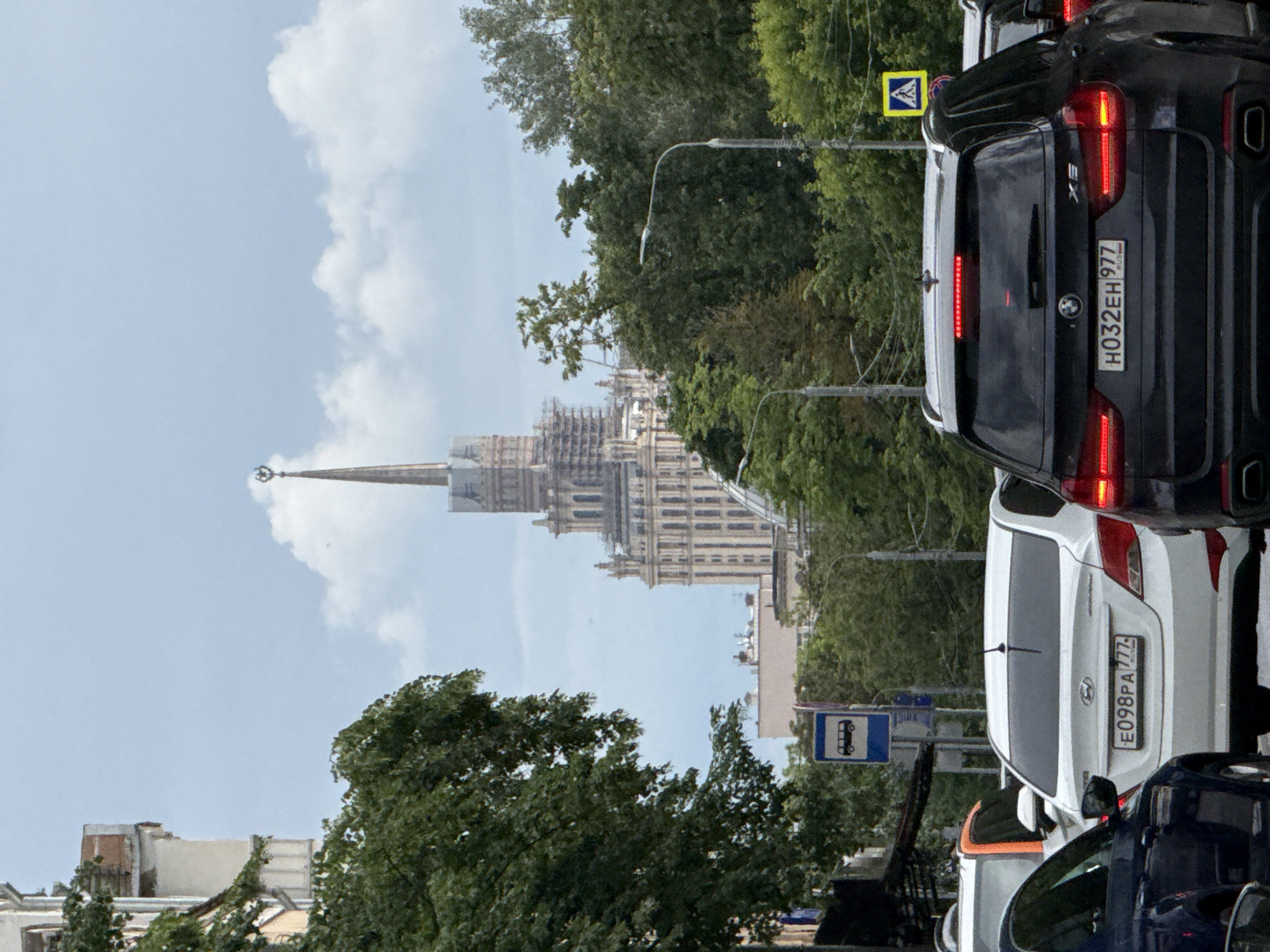 The image shows a city street scene in Moscow, Russia, featuring a traffic jam with several cars. In the background, a prominent Stalinist skyscraper with a spire is visible, undergoing some scaffolding work. The sky is partly cloudy. Among the cars, a black BMW SUV with license plate H032EH 977 is in the foreground. A white Hyundai sedan with license plate E098PA 777 is behind it. Other vehicles and trees line the street, and a bus stop sign is also present.