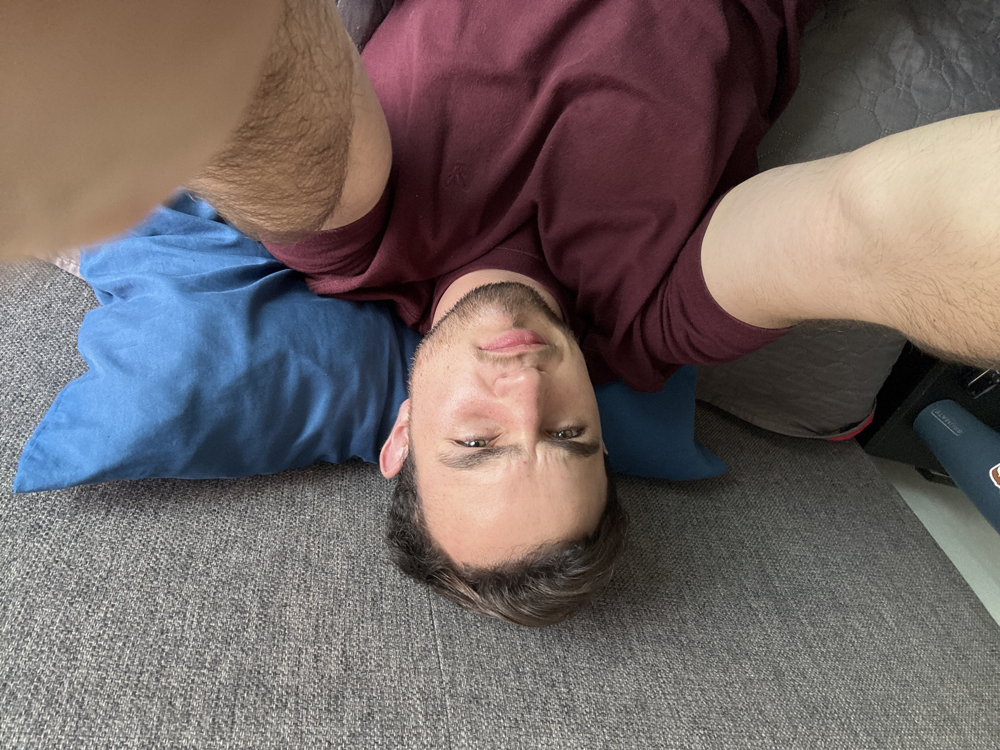A man is lying on a gray couch with a blue pillow under his head. He is wearing a maroon shirt. His arms are raised, one on each side of his head. The photo is taken from an overhead perspective, making the man appear upside down. No information from the image suggests the location is San Bernardino Tlaxcalancingo, Mexico.