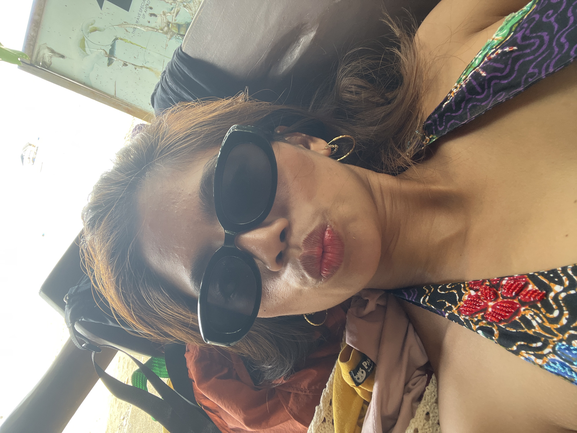 The image shows a person wearing sunglasses and a patterned swimsuit top. They are reclining, possibly on a beach or near water, with various items of clothing and a bag nearby. The location is possibly Banaybanay, Philippines, though without further context, confirmation is impossible.