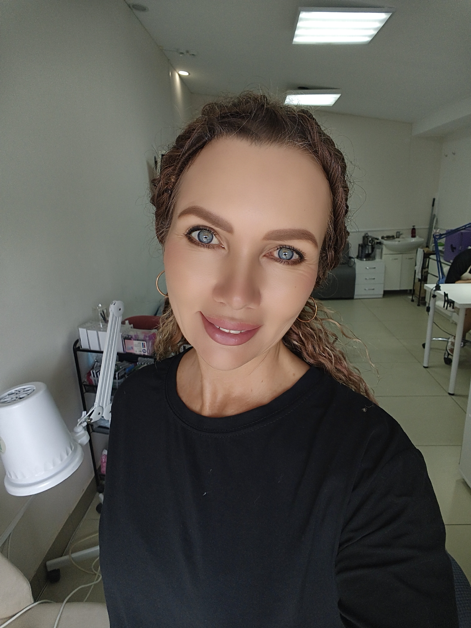 A woman with braided brown hair and blue eyes is taking a selfie in what appears to be a salon or treatment room. She is wearing a black t-shirt and large gold hoop earrings. Behind her, there are shelves stocked with various items, a white sink, and tables with chairs, suggesting a professional setting. The room is brightly lit by overhead fluorescent lights.