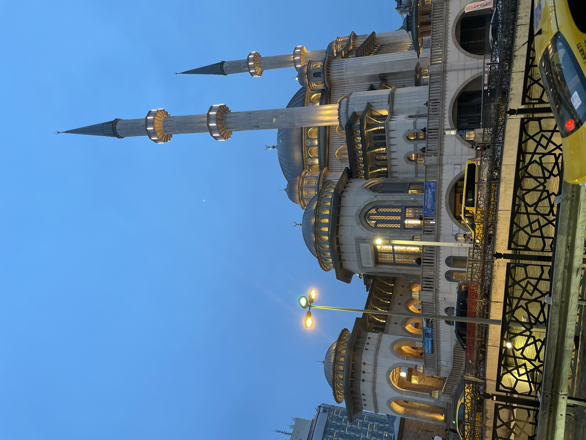 The image depicts a mosque at dusk, possibly in Beyoğlu, Turkey, given the architectural style. The structure is predominantly white with multiple domes and tall, slender minarets illuminated with warm lights. A street lamp is also visible, casting light on the facade of the building. A decorative fence runs along the street. The sky is a deep blue, transitioning into night. A yellow taxi is partially visible on the right side of the frame, suggesting urban traffic. The architectural style of the building and the minarets suggest an Ottoman-era mosque. A modern building can be seen in the background on the lower left.