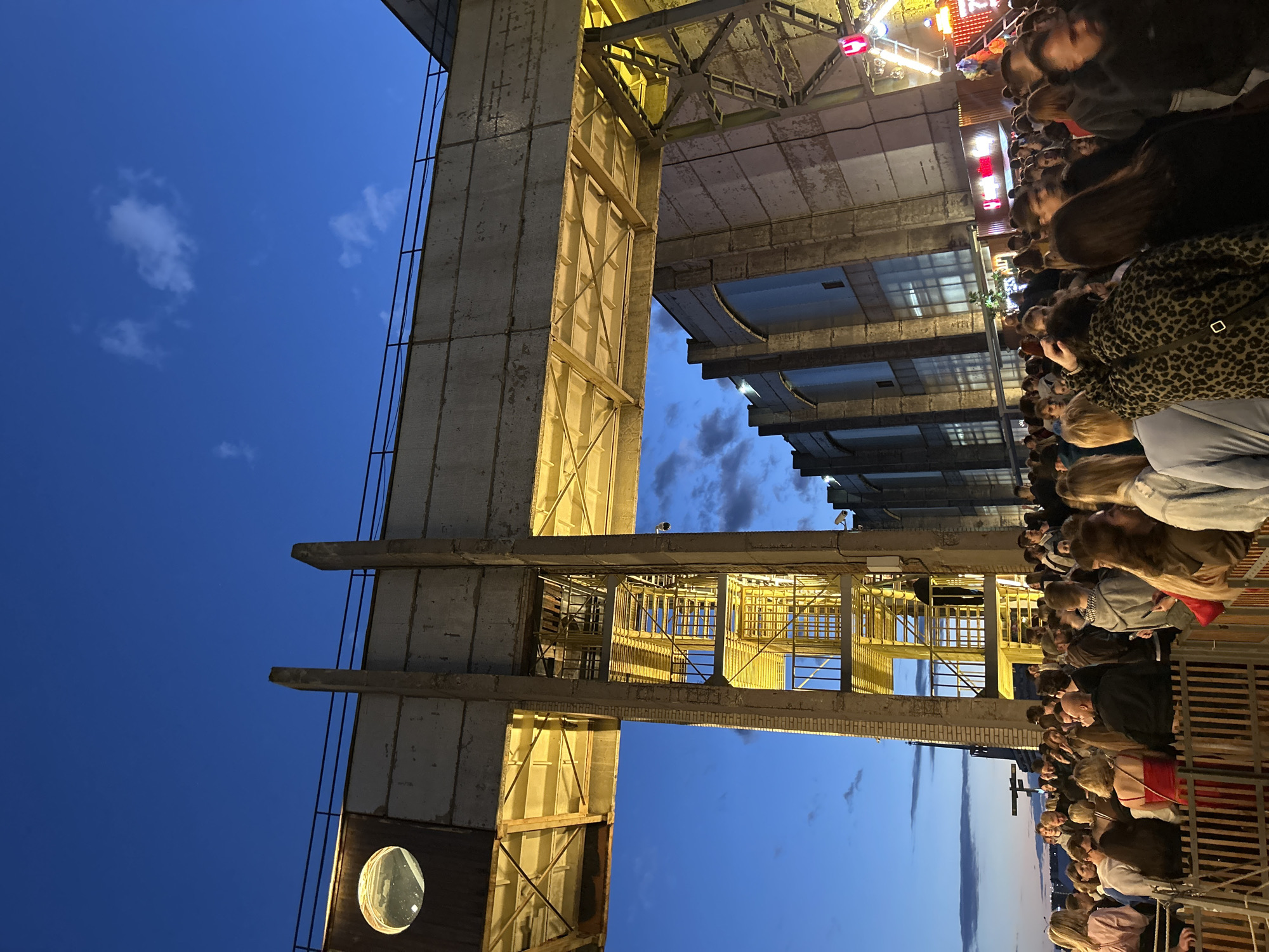 This is a medium shot photograph taken at dusk, capturing a crowd of people gathered outside a building with an industrial aesthetic. The sky is a deep blue with some wispy clouds. A large, elevated concrete structure with yellow metal stairs and walkways spans across the upper portion of the image, partially obscuring the sky. The building itself appears to be a former industrial facility, with large, multi-paned windows and a weathered facade.
In the foreground, a dense crowd of people, dressed in casual attire, are visible. They appear to be waiting or mingling, with some interacting with each other. A wooden fence separates the crowd from the viewer. The lighting suggests an outdoor event or gathering is taking place.
The location is identified as Saint Petersburg, Russia, at the Sevkabel Port, a former cable factory transformed into a public space. The architectural style and the presence of an outdoor gathering are consistent with this location. The overall scene depicts a vibrant evening event in an urban, industrial setting.