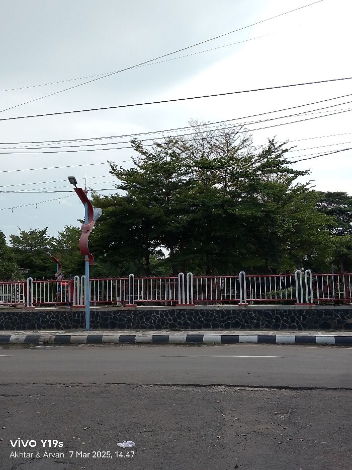 The image shows a street in Indramayu, Indonesia, at the coordinates -6.327 latitude and 108.322 longitude. There is a blacktop road with a white dividing line. The road has a black and white curb and a low stone wall with a red and white metal fence. There are trees behind the fence. In the foreground, there is a small piece of white trash on the road. The sky is overcast. There are many power lines running across the sky. A streetlight stands on the left side of the road. The streetlight is red, white, and blue and has a large curved, red piece at the top. The streetlight is behind the fence. There are no people visible in the scene.