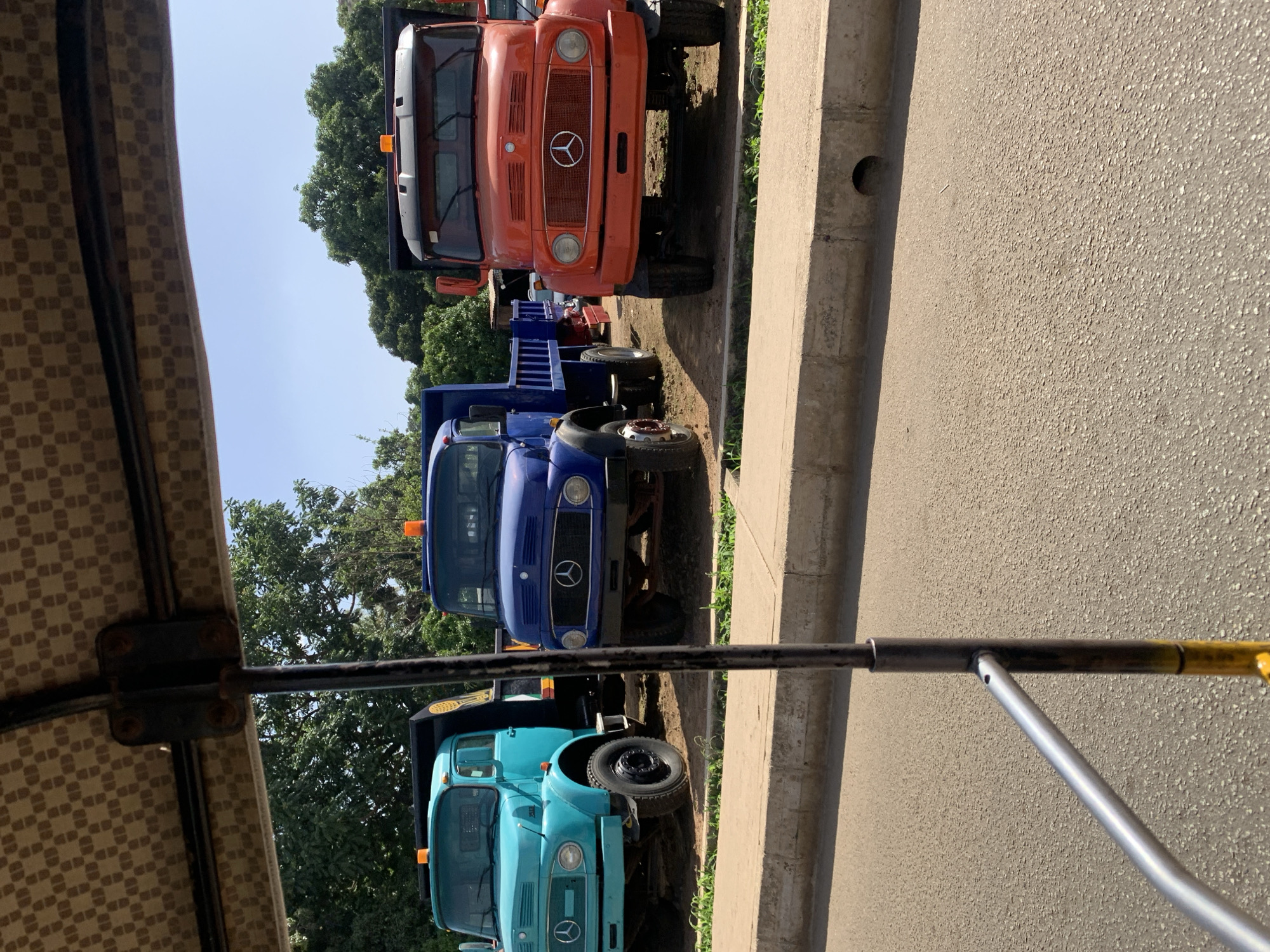 The image shows a view from under a canopy structure, looking out at a row of parked trucks. The nearest truck is light blue, followed by a blue truck, and then an orange truck. All three trucks have the Mercedes-Benz logo on their front grilles. The trucks are parked on the side of a paved road, next to a concrete curb. Trees are visible in the background. The location is likely near the New Stadium in Nigeria, given the presence of older-model Mercedes-Benz trucks often used for commercial purposes in African countries. No people are visible in the image.