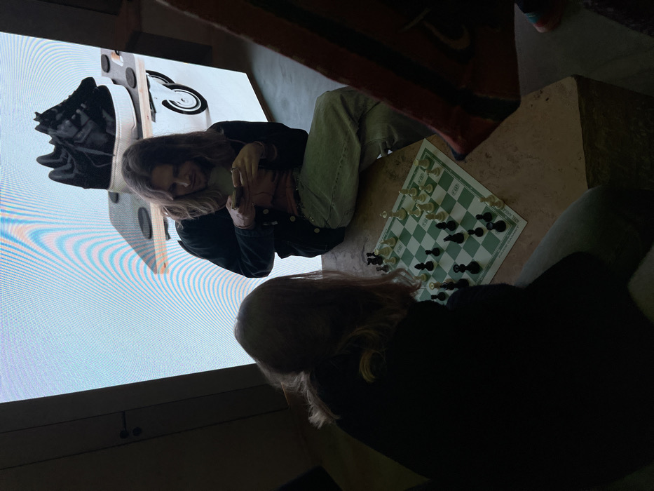 Two people are engaged in a game of chess on a green and white checkered board resting on a stone surface. One person, facing away from the camera, wears a black shirt. The other person, facing a large screen displaying a shoe, wears a denim jacket and light-colored pants. Part of a patterned rug is visible above the chessboard.