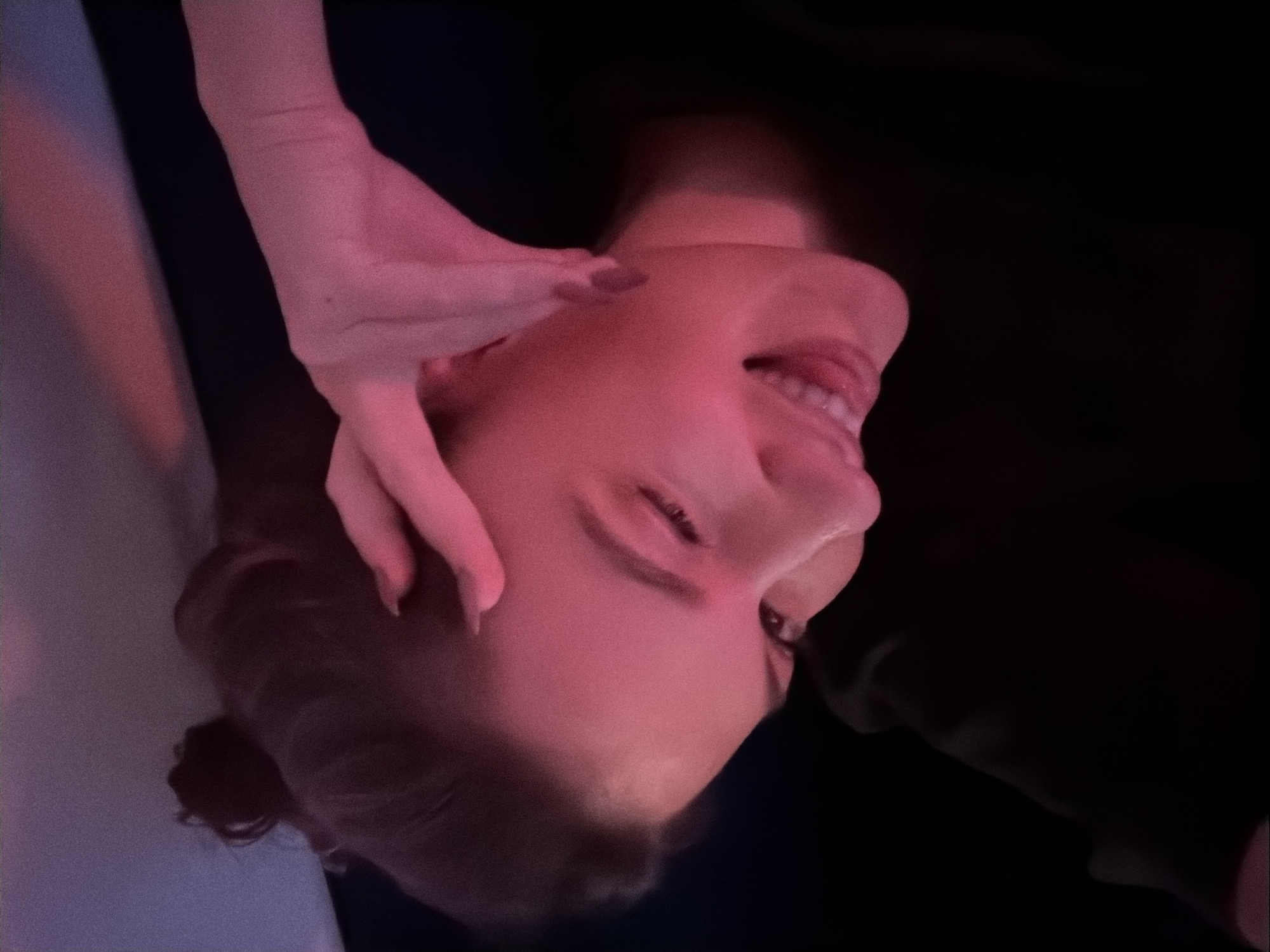 A person's face is seen close up, tilted at an unusual angle, with a hand on their face near the ear. The skin is illuminated with a pinkish or reddish light. The background is mostly dark, with hints of fabric or furniture. The setting cannot be identified as being in Belgrade, Serbia, based on visual information alone.