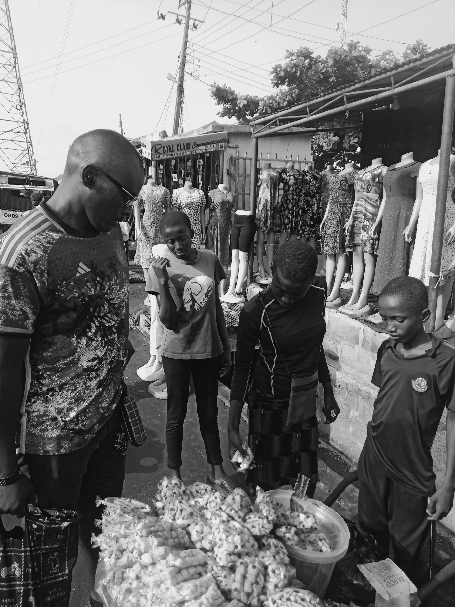 The black and white photograph depicts a street scene in Onitsha, Nigeria, where a group of individuals appear to be engaged in a transaction. An adult male, wearing glasses and a patterned Adidas shirt, stands to the left, looking down at a display of what appears to be pre-packaged food items or snacks. Next to him is a younger person, wearing a t-shirt with a graphic design. Two more children stand to the right of the food display, their attention directed towards it. The environment suggests an outdoor market setting, with a clothing store visible in the background, featuring mannequins dressed in various dresses. The store sign reads "ROYAL CLASS Communications, DEALERS ONINSTALLING OF PHONE ACCESSORISMI". A tower is also seen in the background. The presence of shopping bags with the word "AFRICA" on them, along with the "RASCO" snack bag, adds further details to the scene's context.