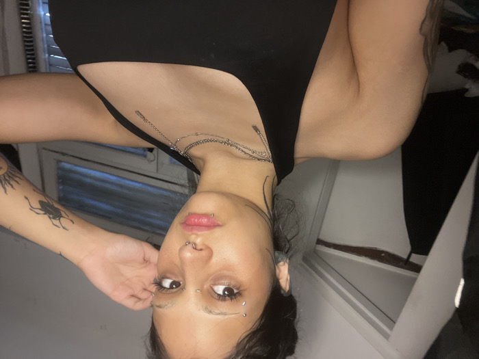 A person with piercings and tattoos is angled upside down. They wear a black top and several silver chain necklaces. A large insect tattoo and a sun tattoo are visible on the left arm. The person has piercings on the lips, nose, and eyebrows. A window with blinds is in the background on the left. A white wall and a portion of a black garment are visible on the right.